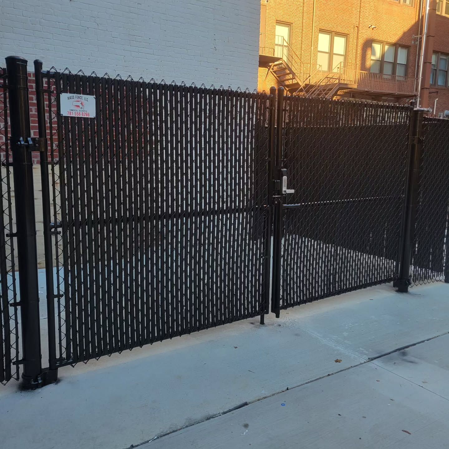 Black chain-link fence with privacy slats and a gate along a sidewalk, with a building in the background.