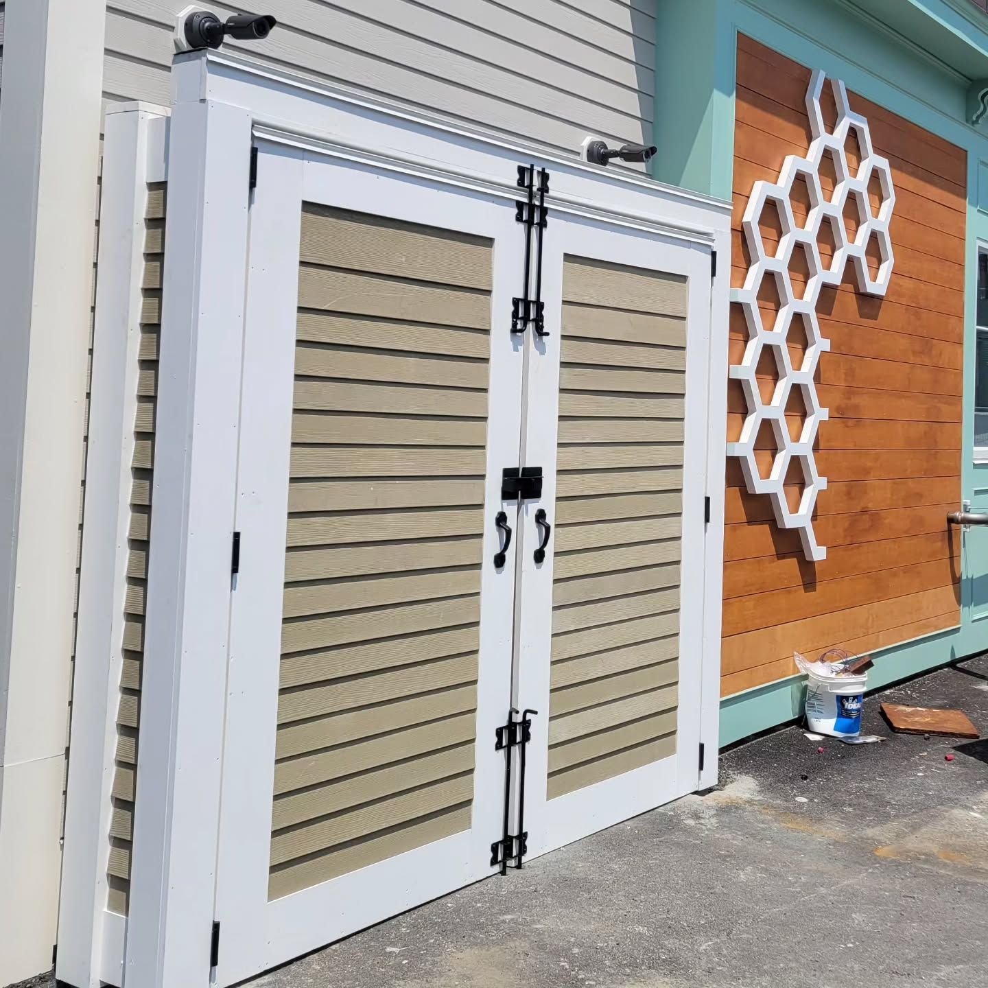 White framed double doors with tan horizontal slats and black hardware, near a wooden wall with a honeycomb design.