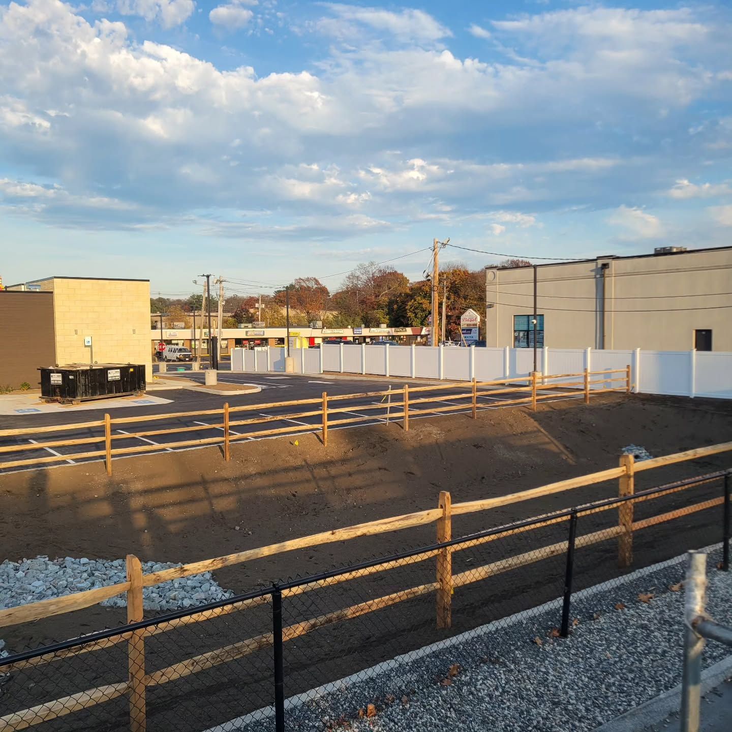 Construction site with wooden fence, white fence, and buildings under a cloudy sky.