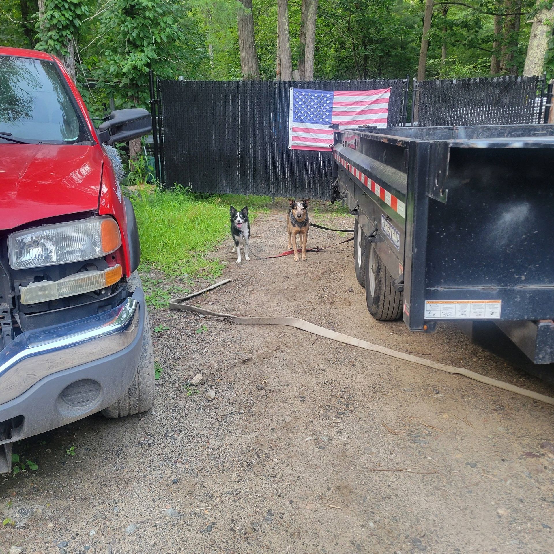 Red truck, black trailer, two dogs, American flag on a black fence in a dirt lot.
