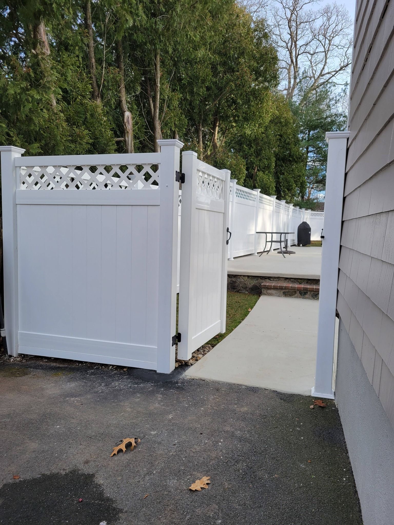White vinyl fence with gate, concrete walkway, and house siding in a yard with trees.