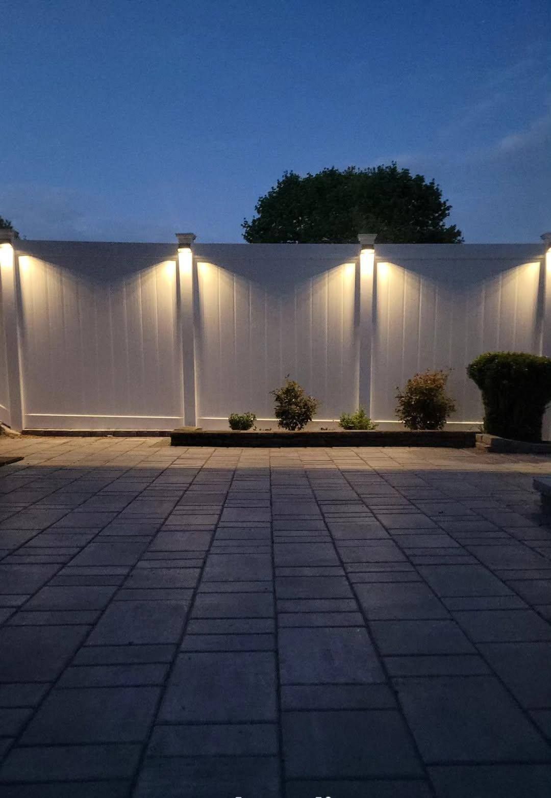 White fence with built-in lights illuminates a dark patio and small plants at dusk.