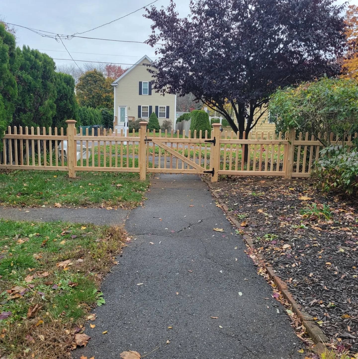 A light-colored wooden picket fence with a gate surrounds the front yard of a house on a cloudy day.