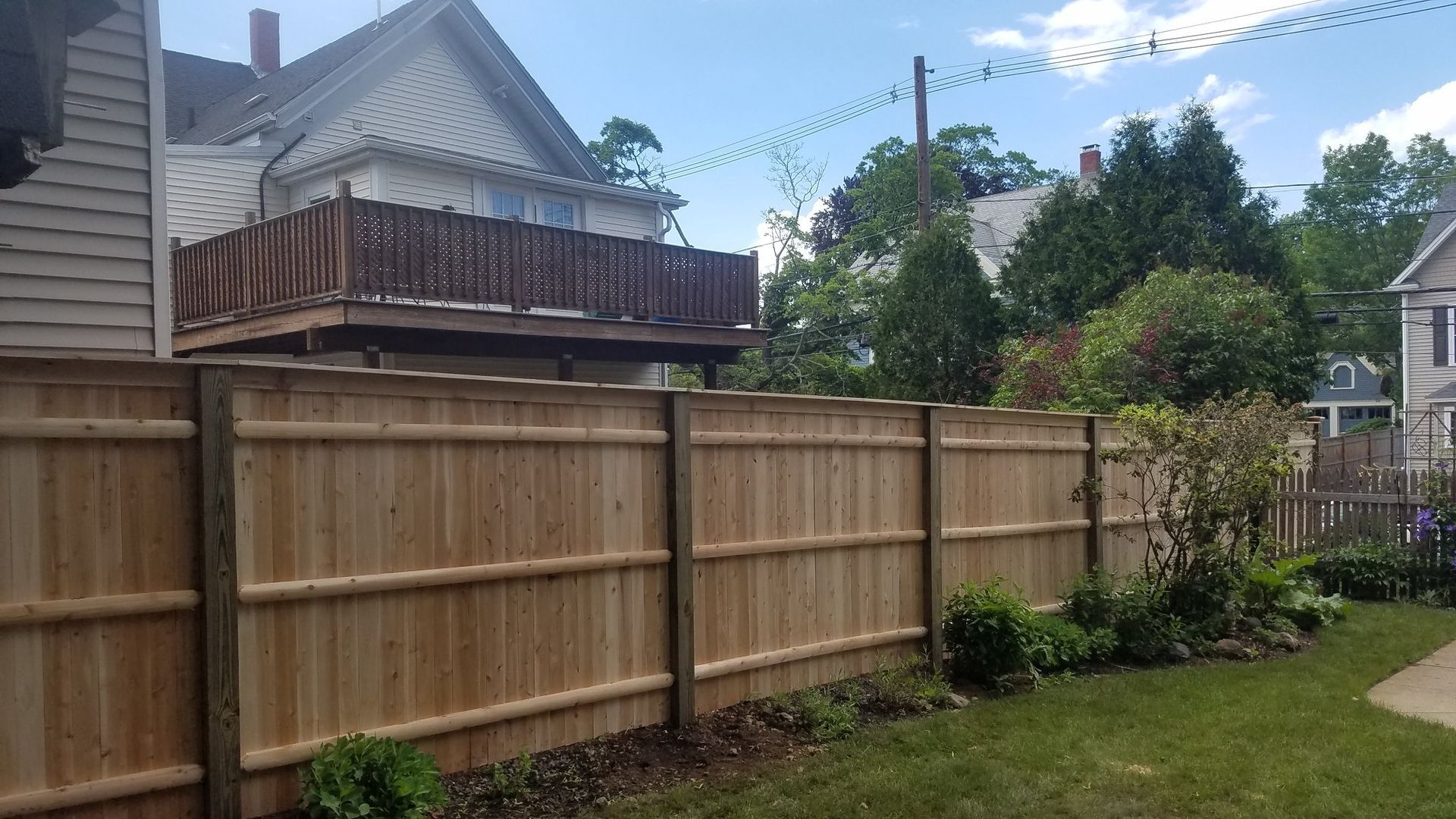 Wooden fence in a backyard; a house with a deck is in the background, sunny day.