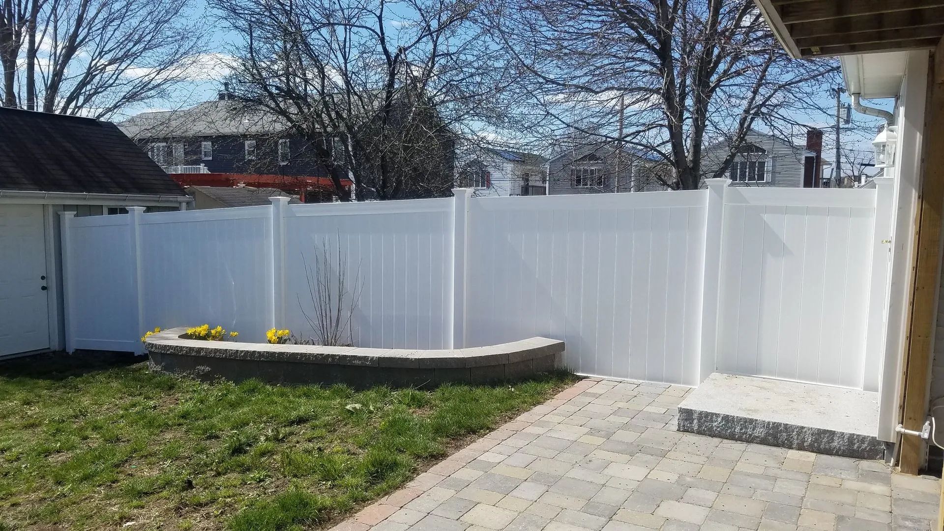White vinyl fence around a backyard with a raised garden bed, grass, and stone patio in sunlight