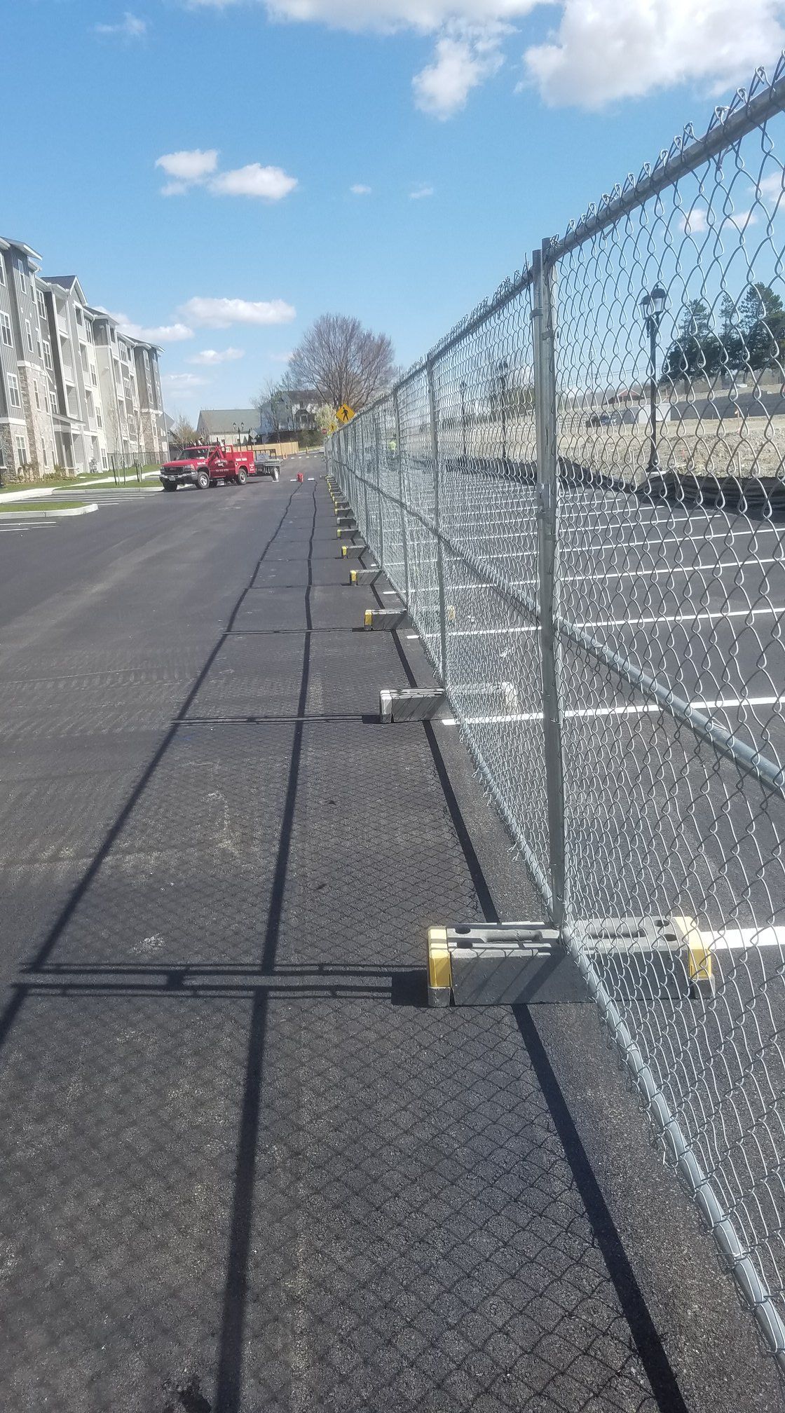 Chain-link fence along new asphalt parking lot, blue sky, parked cars and apartment building in the background.
