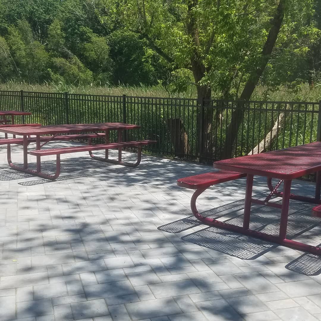 Red picnic tables on a paved area, near a fence and trees. Sunlight creates shadows.
