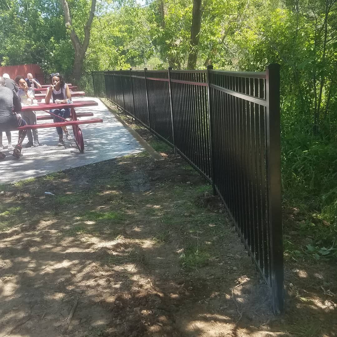 Black metal fence along a dirt and concrete area with trees and people in the background.