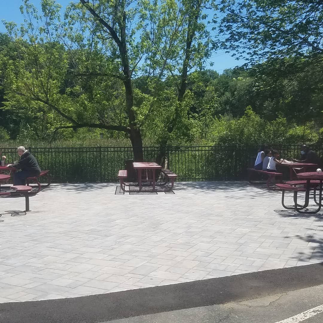 Picnic tables on a gravel patio under trees. People sit at some tables. Green foliage and fence in background.
