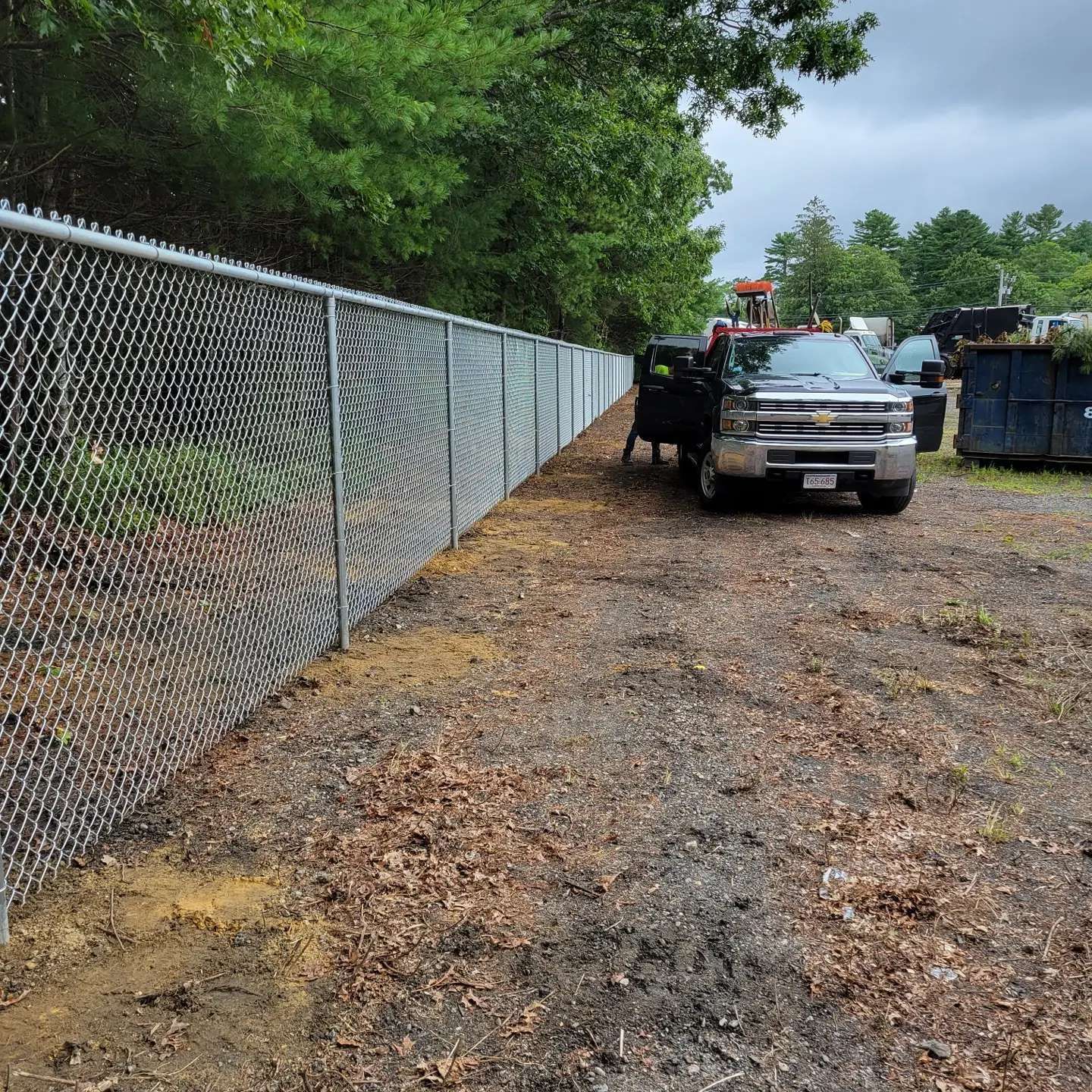Chain-link fence beside a dirt path with a parked truck; trees and dumpsters in the background.