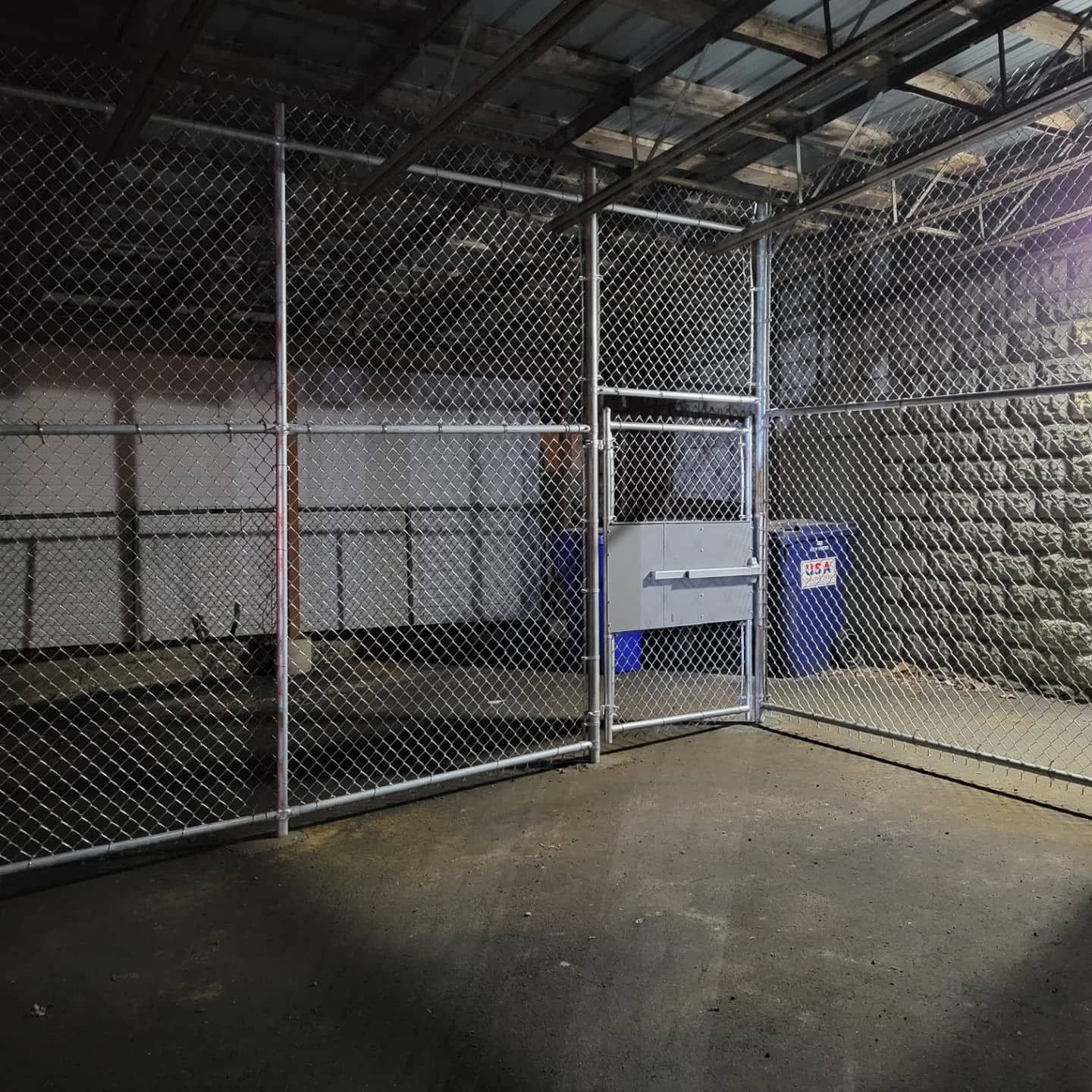 Chain-link fenced enclosure with a gate. Concrete floor, brick wall on right, and blue trash can visible.