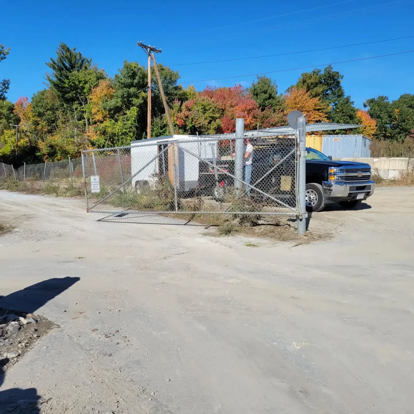 A truck exiting a gated area with a trailer on a dirt road, trees with fall foliage in the background.