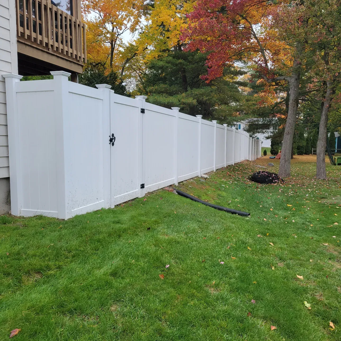 A white vinyl privacy fence with a matching gate extends from the side of a house into a grassy, tree-lined yard.