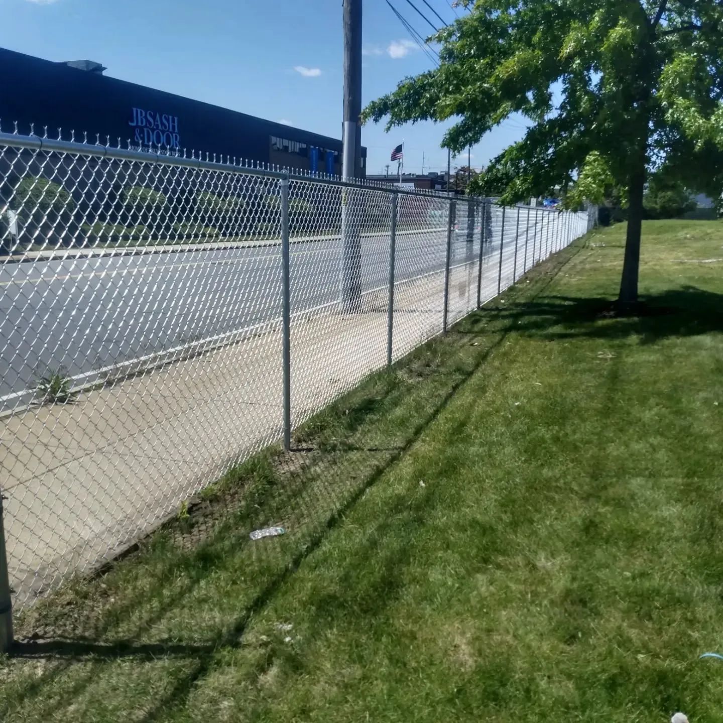 Chain-link fence along a sidewalk beside a grassy strip and road on a sunny day