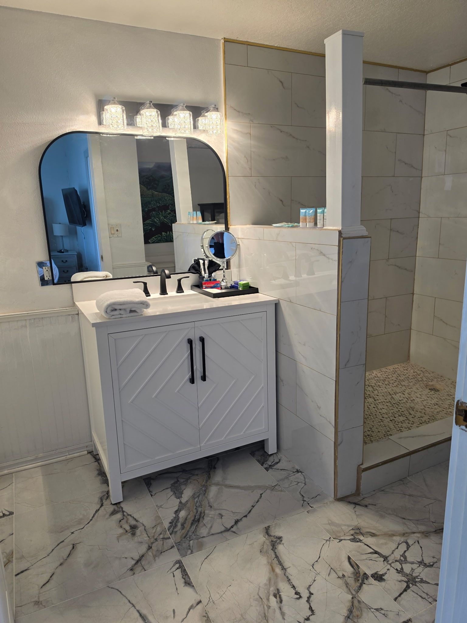 Bathroom vanity with mirror, lights, and shower stall; white cabinet and marble-patterned tile floor.