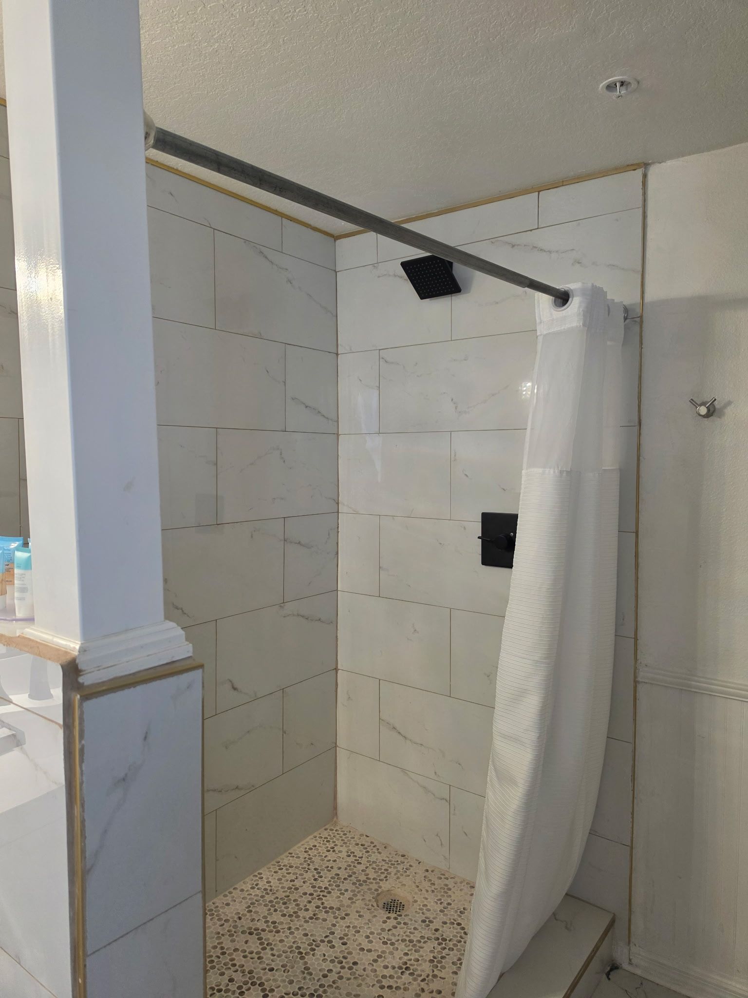Small tiled shower stall with a partially open white shower curtain and pebble-style floor.