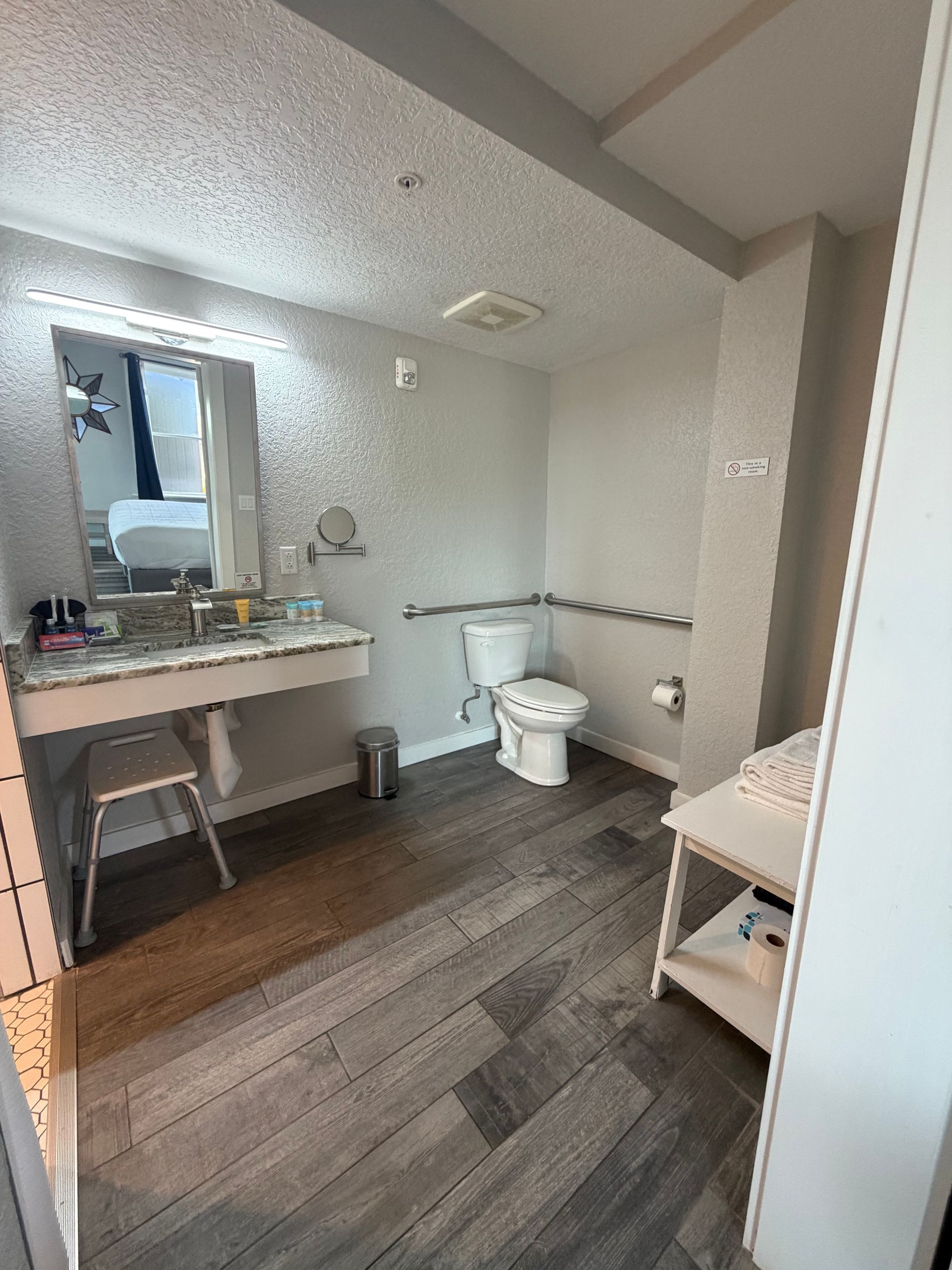 Small accessible bathroom with sink, mirror, toilet, grab bars, and dark wood flooring.