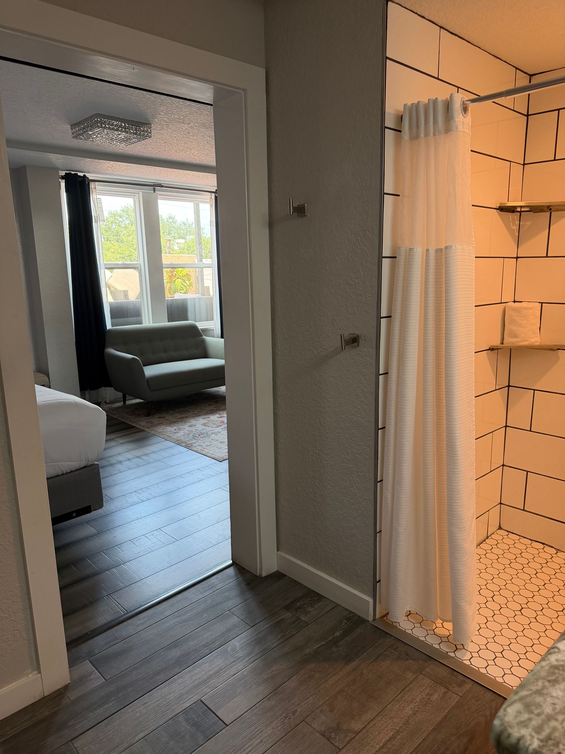 Hallway opening to a bedroom and sunlit living area, with a shower stall on the right.