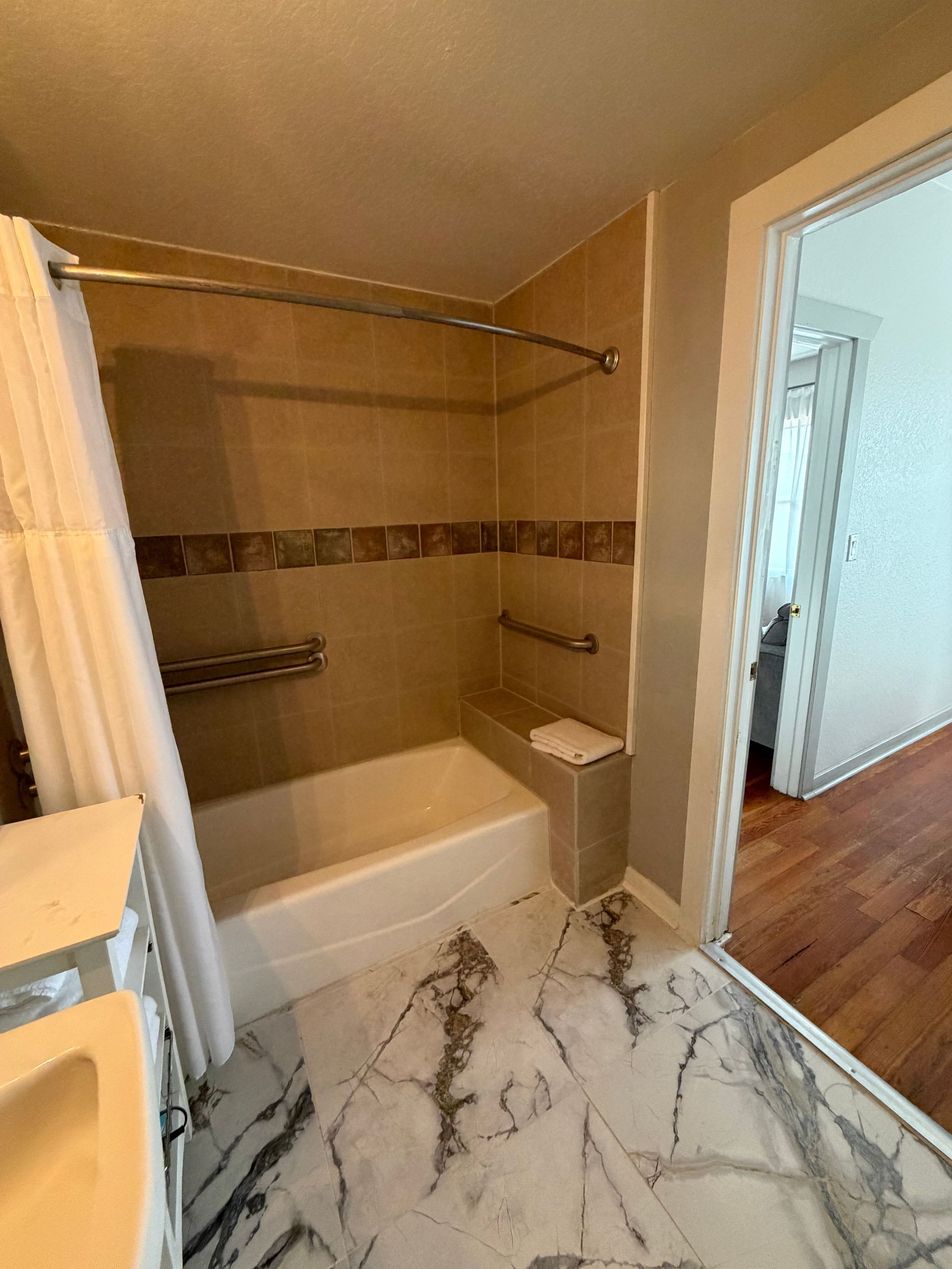 Small bathroom with a white tub, beige walls, marble-pattern floor, and open doorway to a hallway