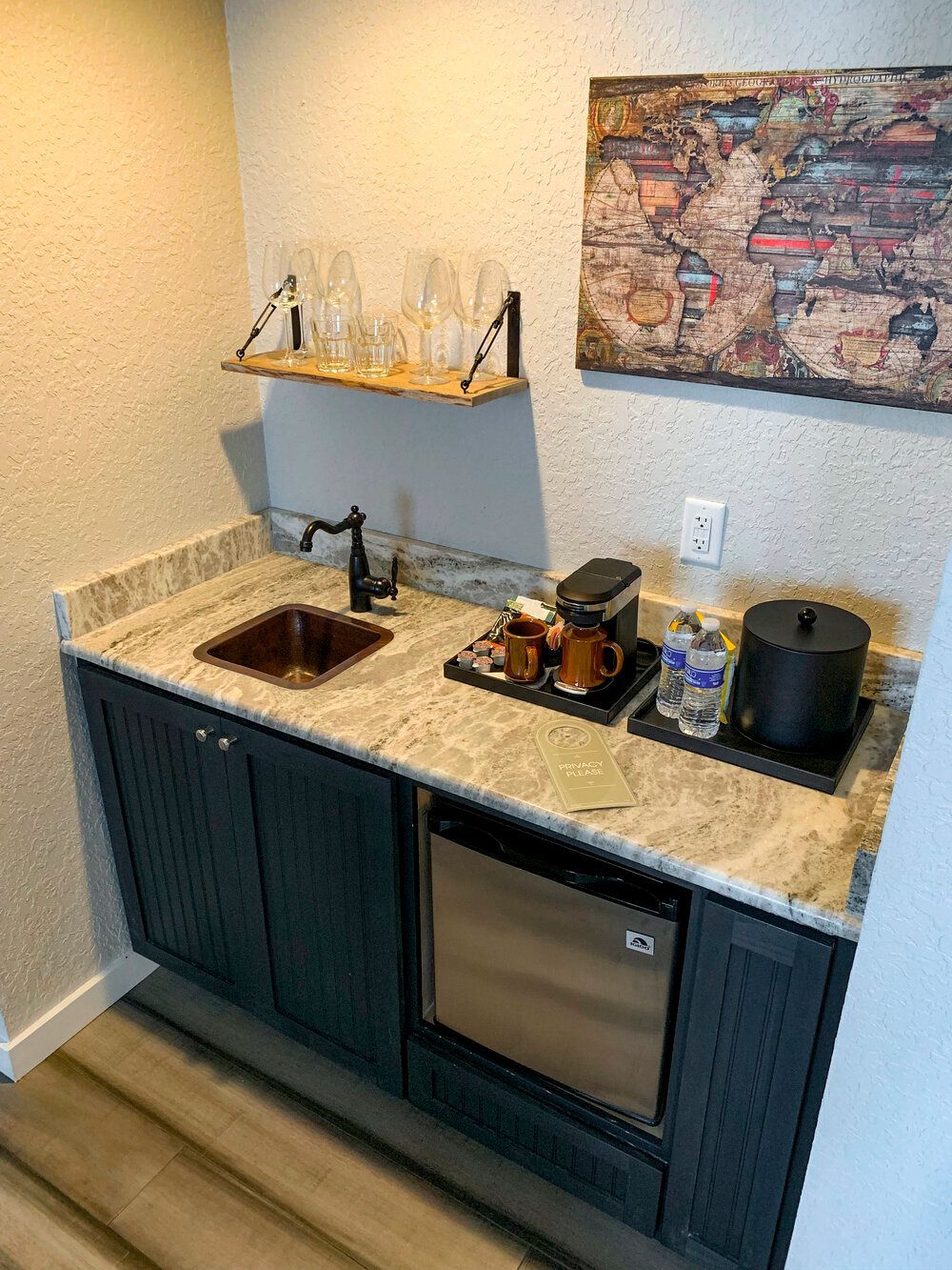Compact home bar with granite countertop, sink, mini fridge, and dark cabinets beneath wall art.