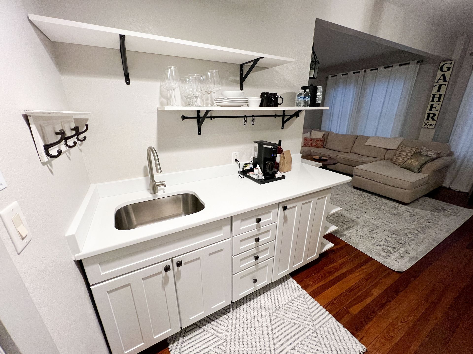 White kitchenette with sink, shelves, coffee maker, and white cabinets beside a living room.