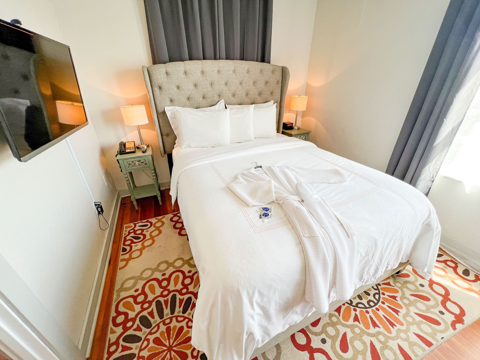 Bright bedroom with a neatly made white bed, tufted headboard, bedside lamps, wall TV, and patterned rug.