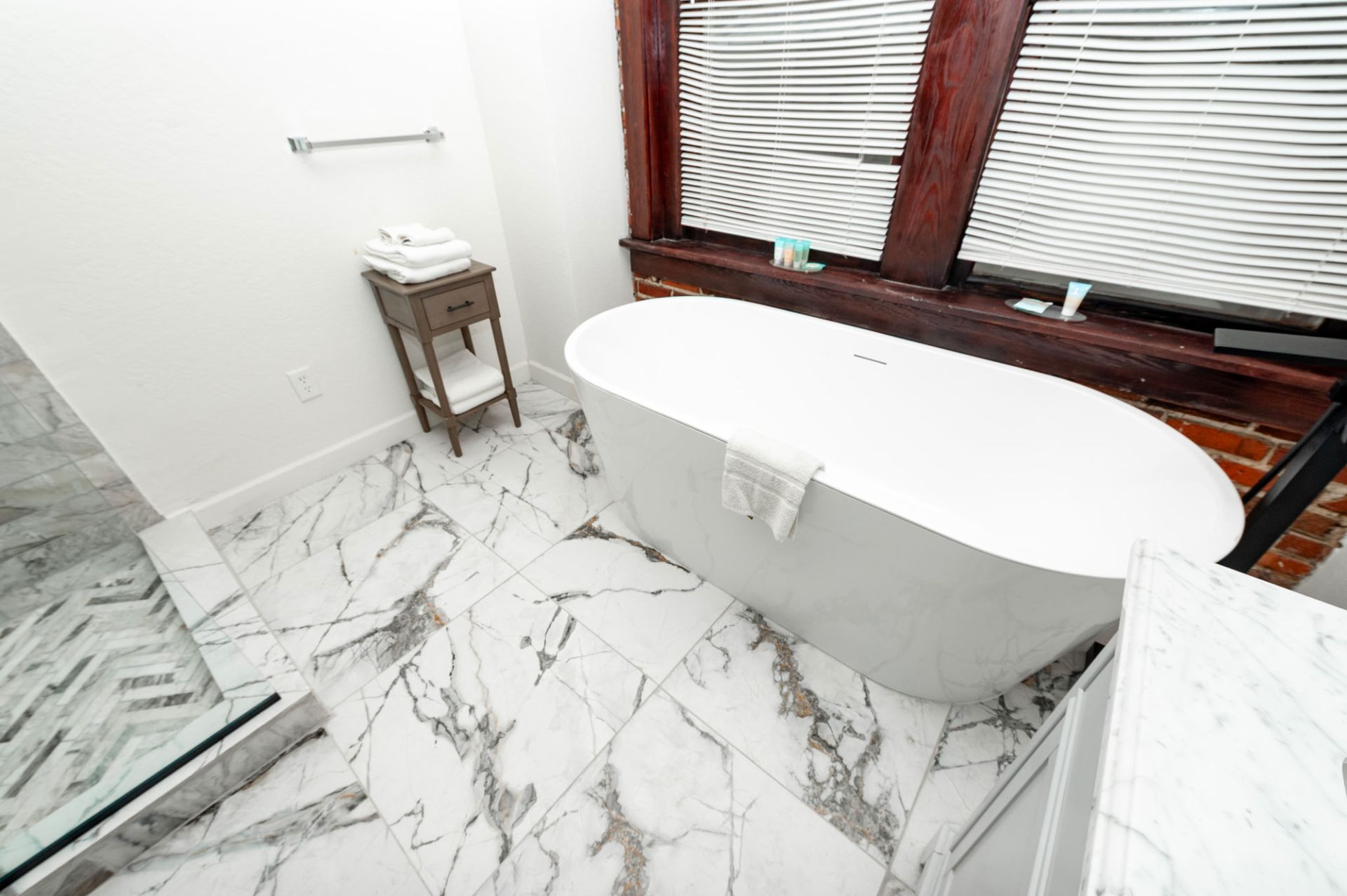 Modern bathroom with freestanding white tub, marble tile floor, and wood-trimmed windows with blinds