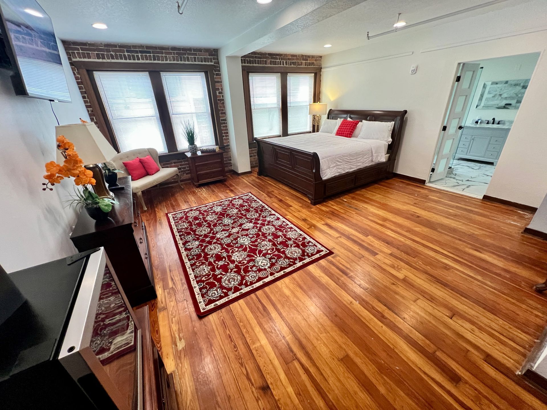 Bright bedroom with hardwood floor, red rug, gray bed, and large windows with blinds