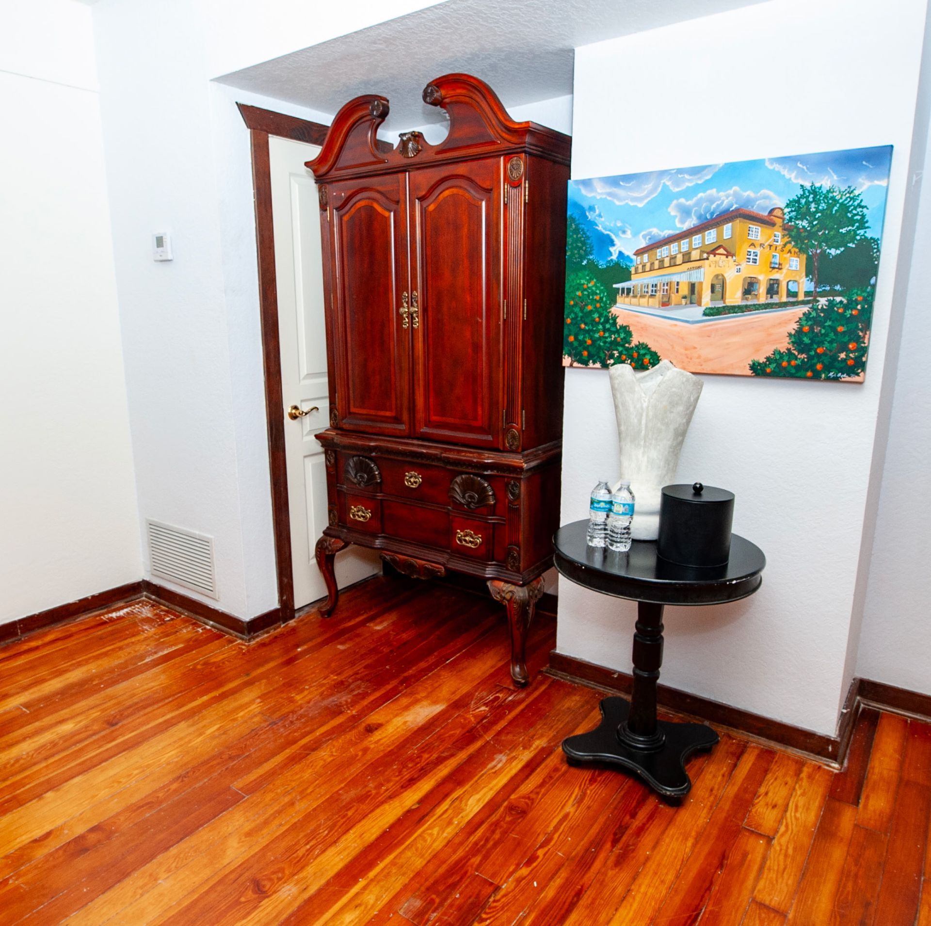 Bright room with polished wood floor, ornate wooden cabinet, small round table, and colorful wall painting.