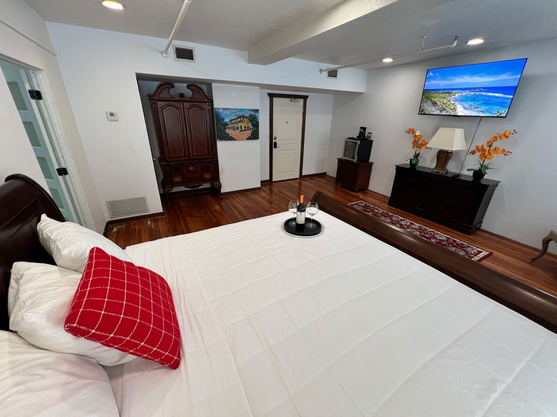 Bedroom with white bedspread and red pillow, dark wood furniture, wall TV, and fireplace in a bright room