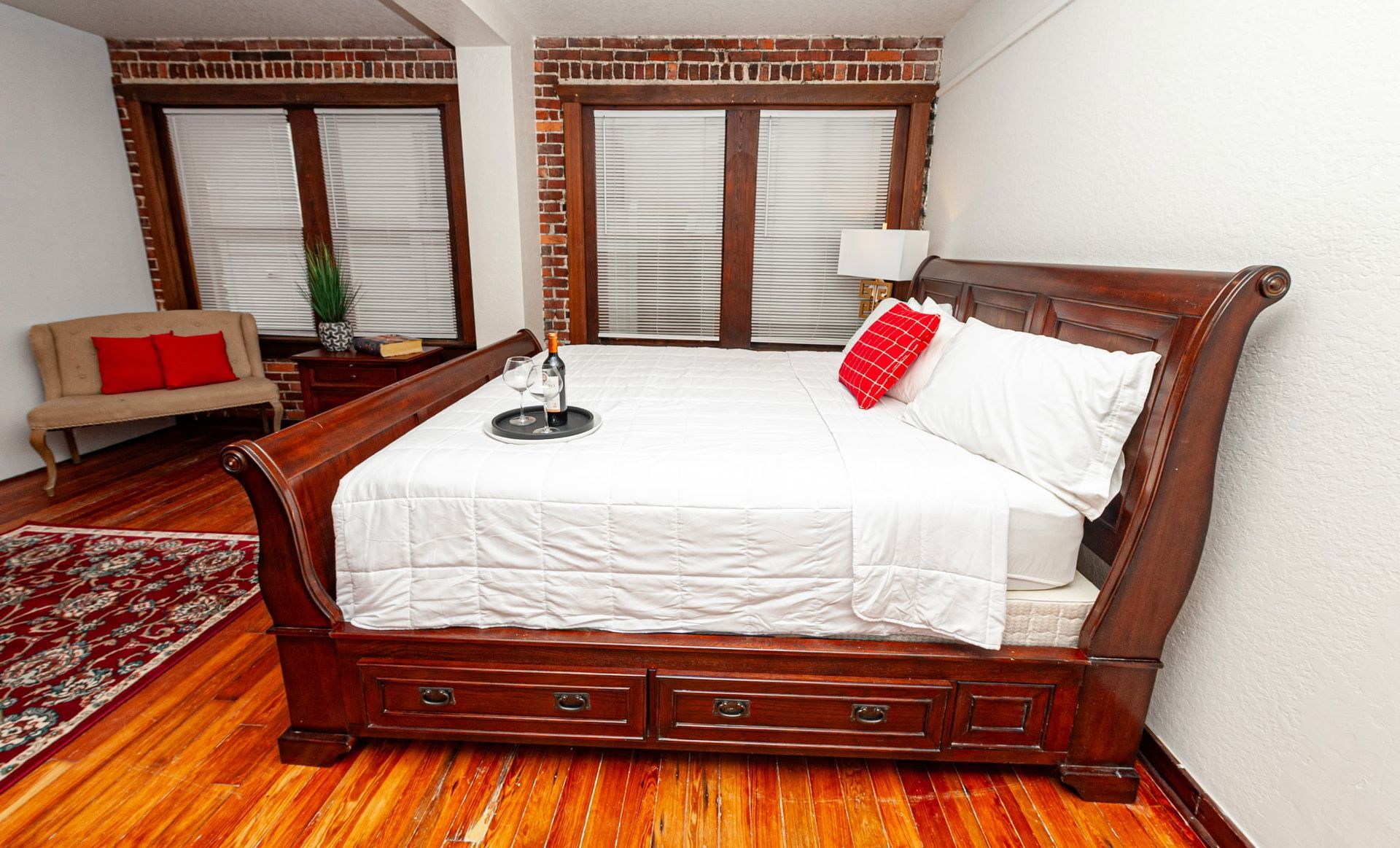 Wooden bedroom with a king bed, white bedding, red accent pillow, windows, and patterned rug