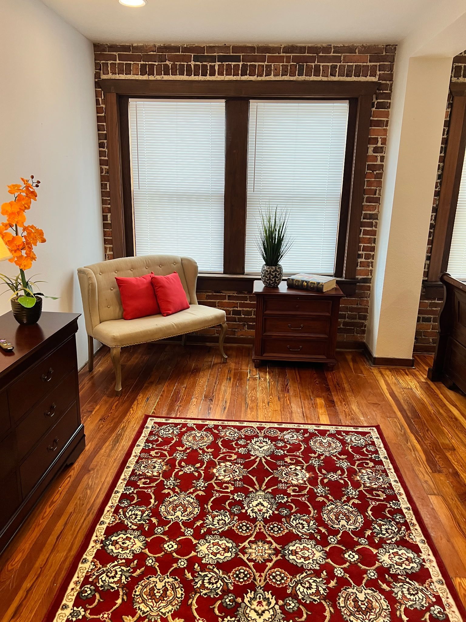 Cozy living room with red patterned rug, tan chair, wooden dresser, and frosted windows.