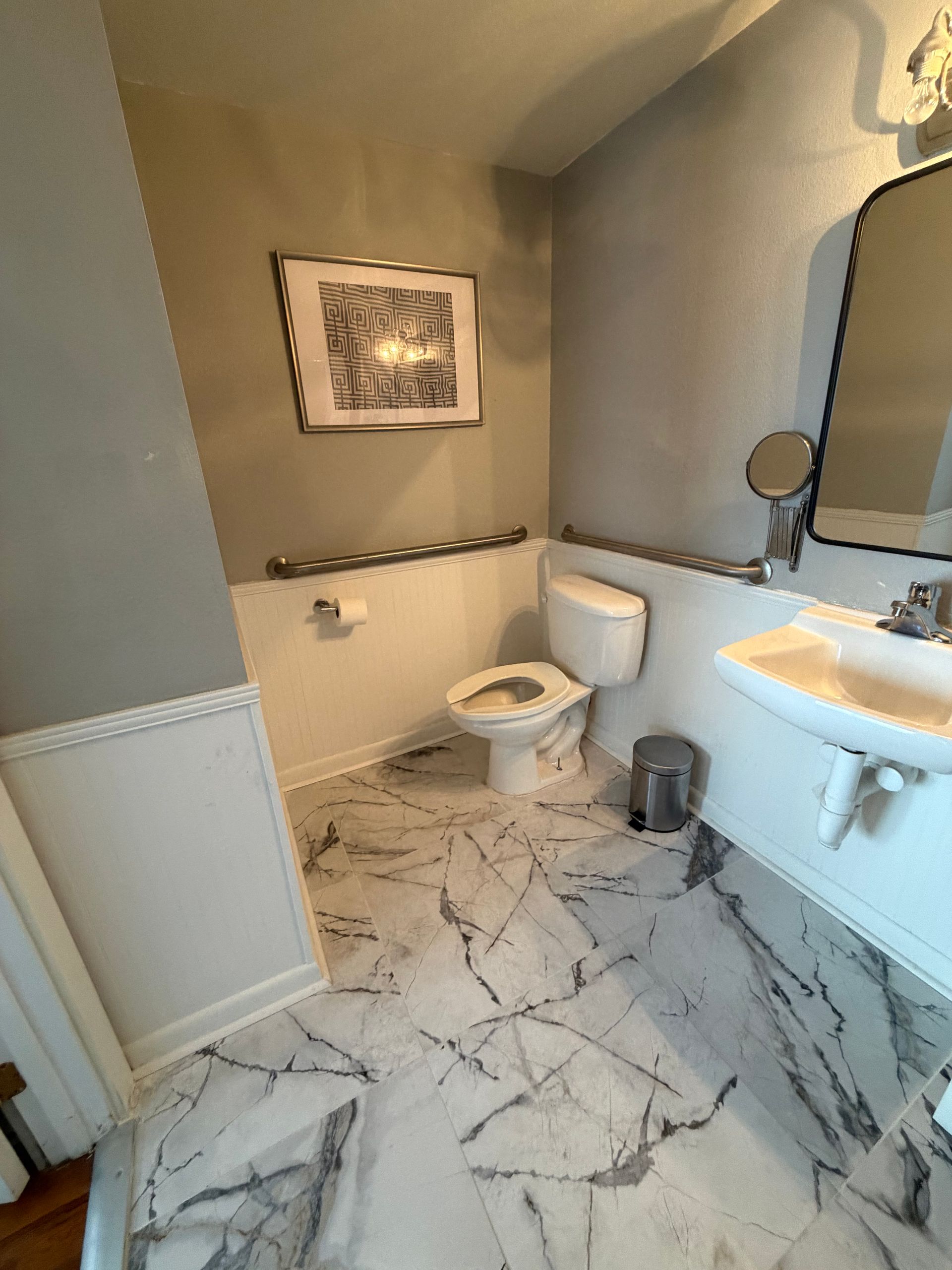 Small bathroom with a toilet, sink, mirror, gray walls, and marble-pattern tile floor.