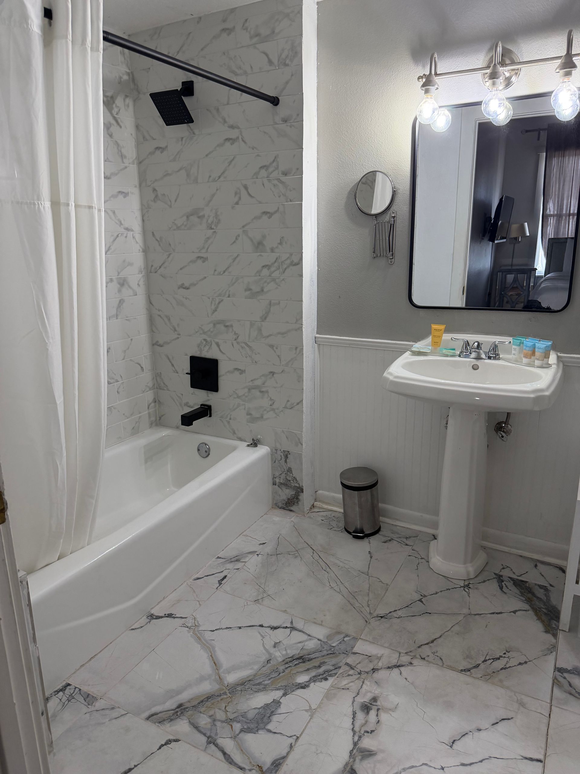 Small white marble bathroom with tub, pedestal sink, mirror, and shower curtain