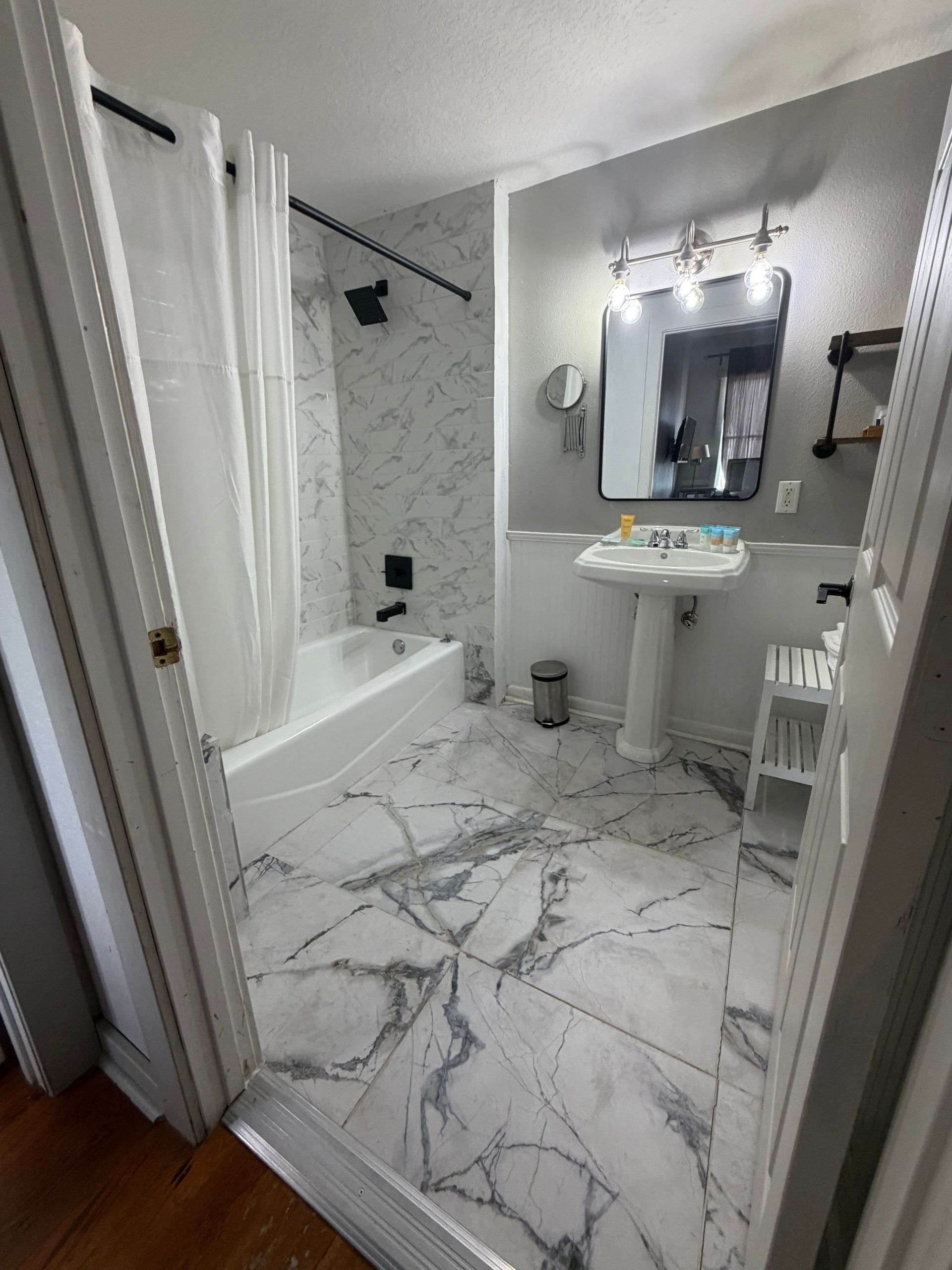 Small white bathroom with marble floor, bathtub, pedestal sink, mirror, and shower curtain.