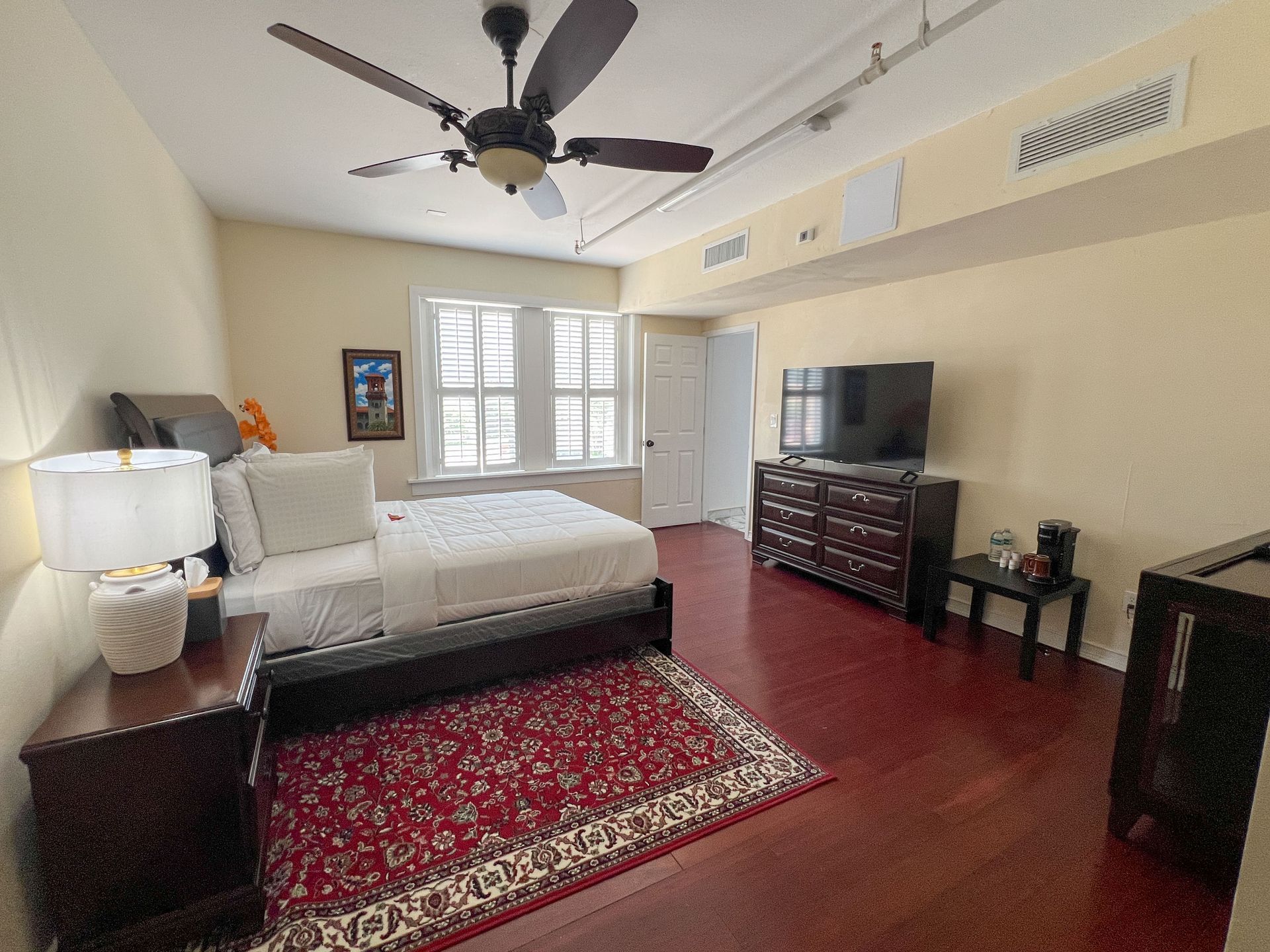 Spacious bedroom with a bed, red rug, ceiling fan, dresser, TV, and white French doors.