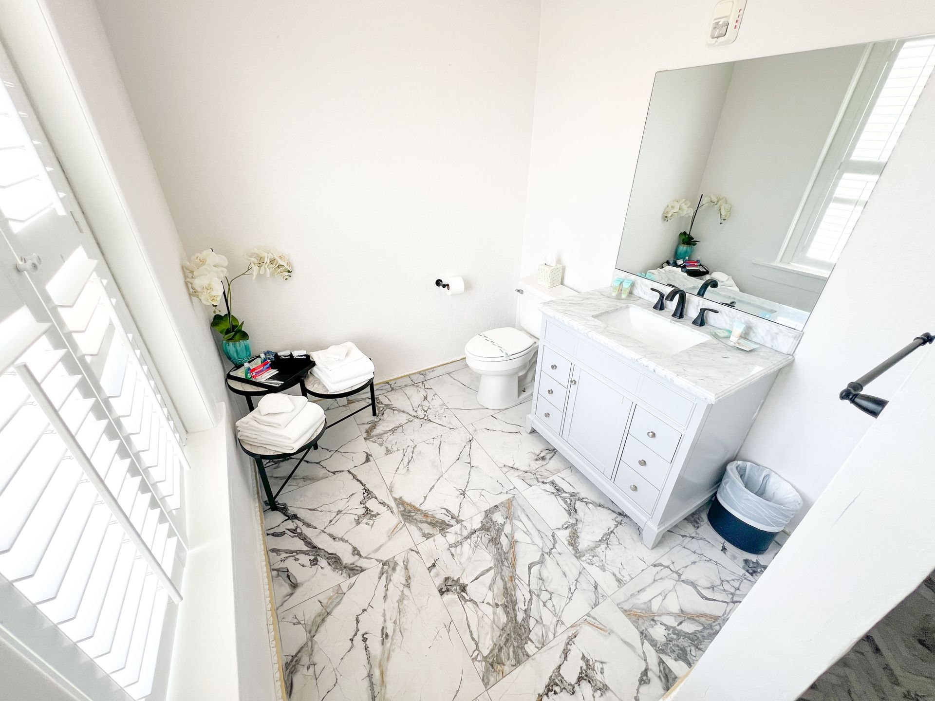 Bright white bathroom with marble-pattern tile, vanity sink, mirror, toilet, and windows with shutters
