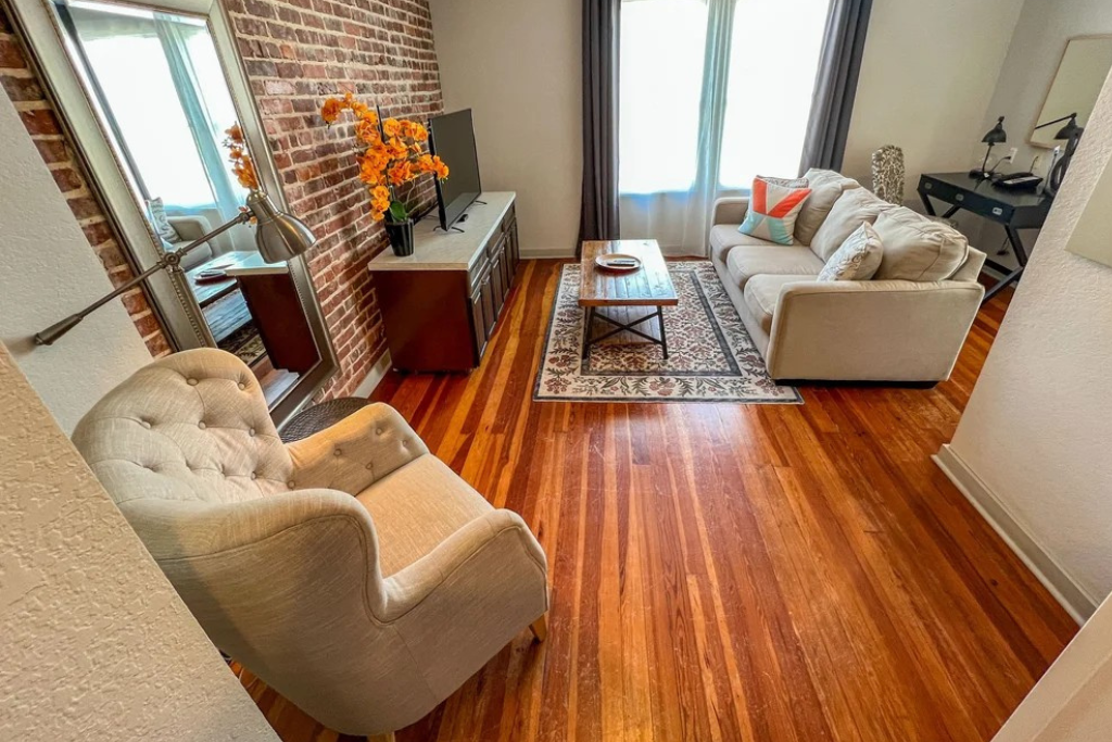 Bright living room with wood floors, tufted chairs, sofa, rug, and brick accent wall