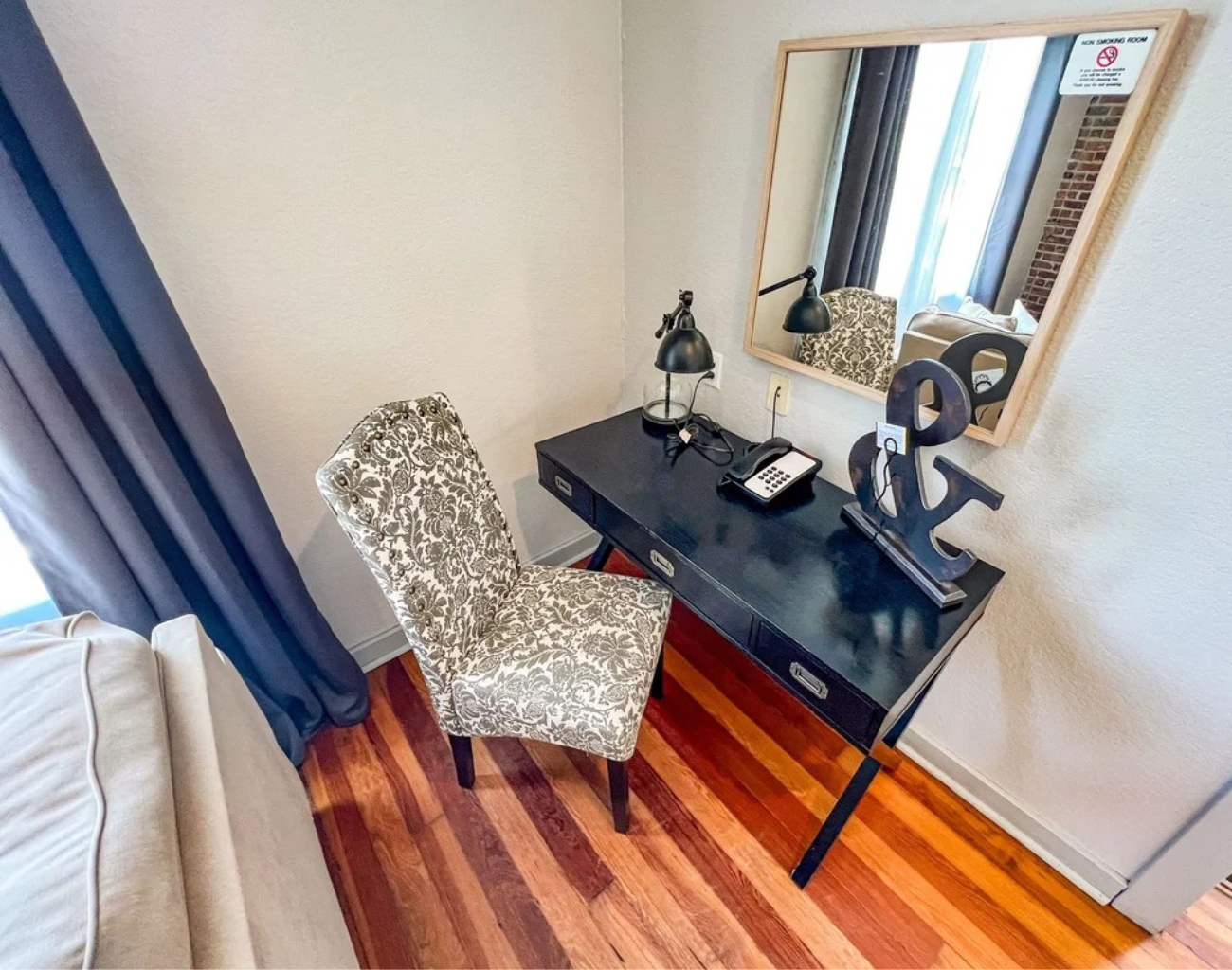 Small home office with patterned chair, black desk, lamp, mirror, and window with blue curtains on wood floor.