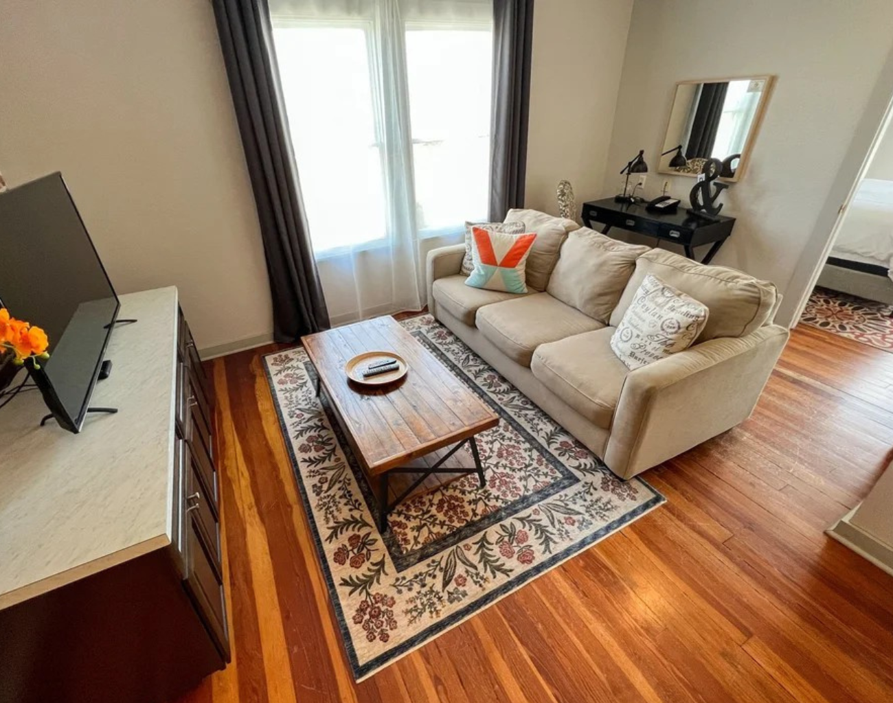 Bright living room with beige sofa, coffee table, patterned rug, and hardwood floors by a window.