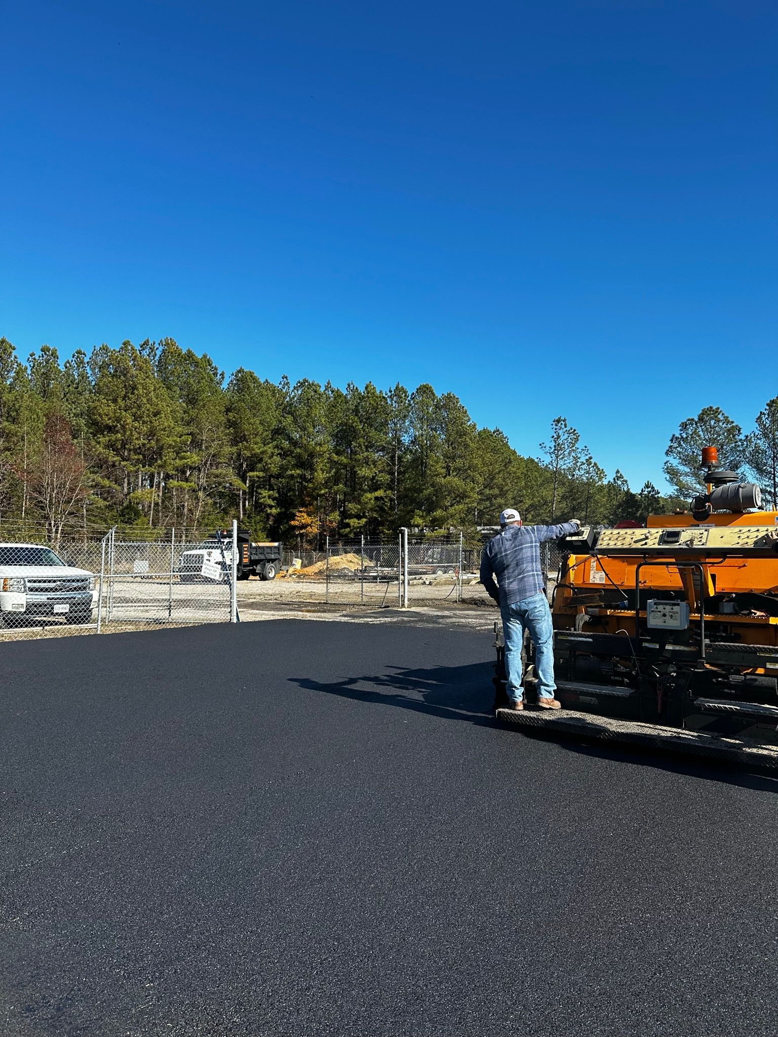 Worker standing beside orange road paver on a newly paved black asphalt lot under a clear blue sky