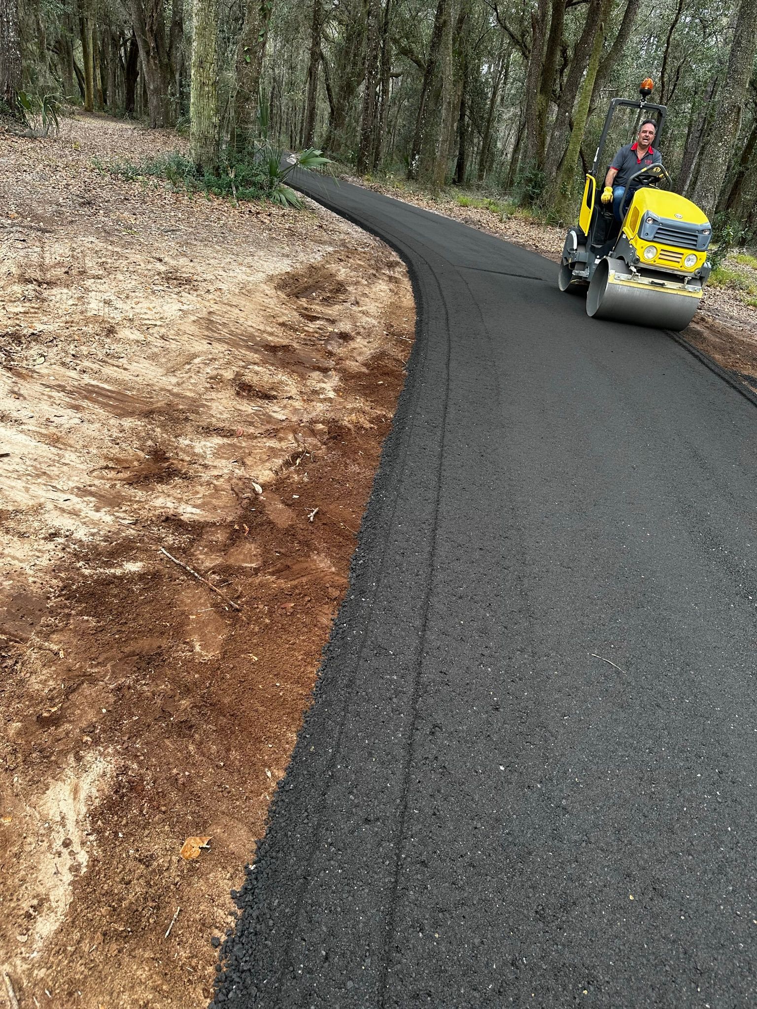 Yellow road roller paving a curving forest path beside leaf-covered ground.
