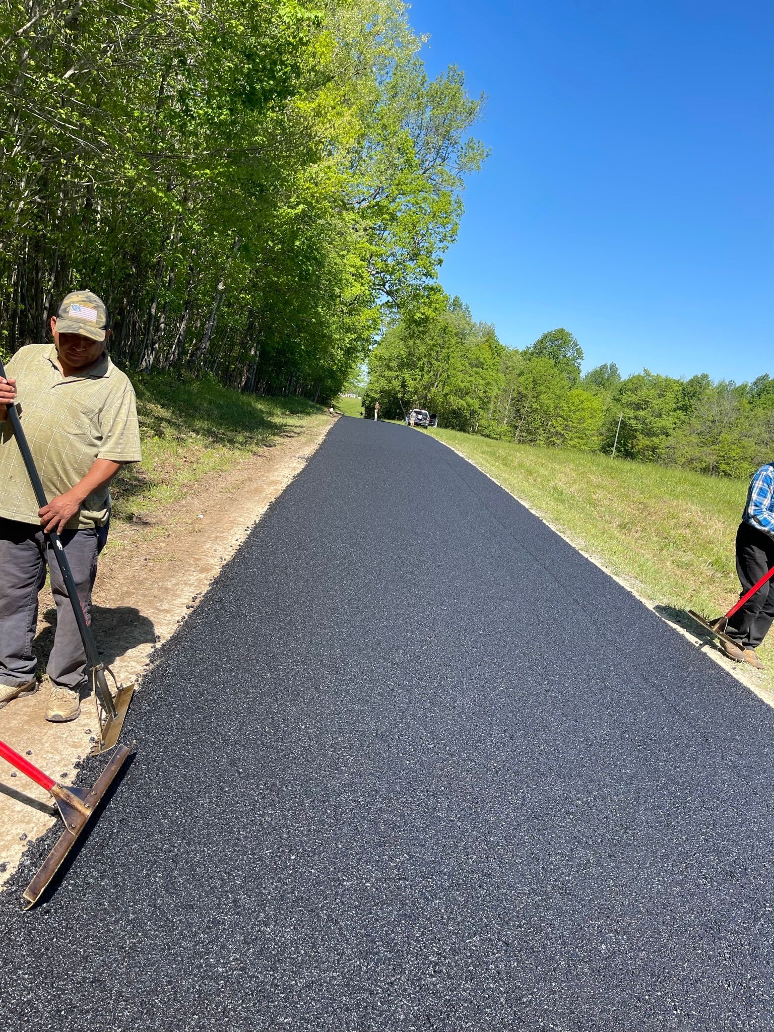 Workers paving a fresh black asphalt road beside a wooded roadside under a clear blue sky