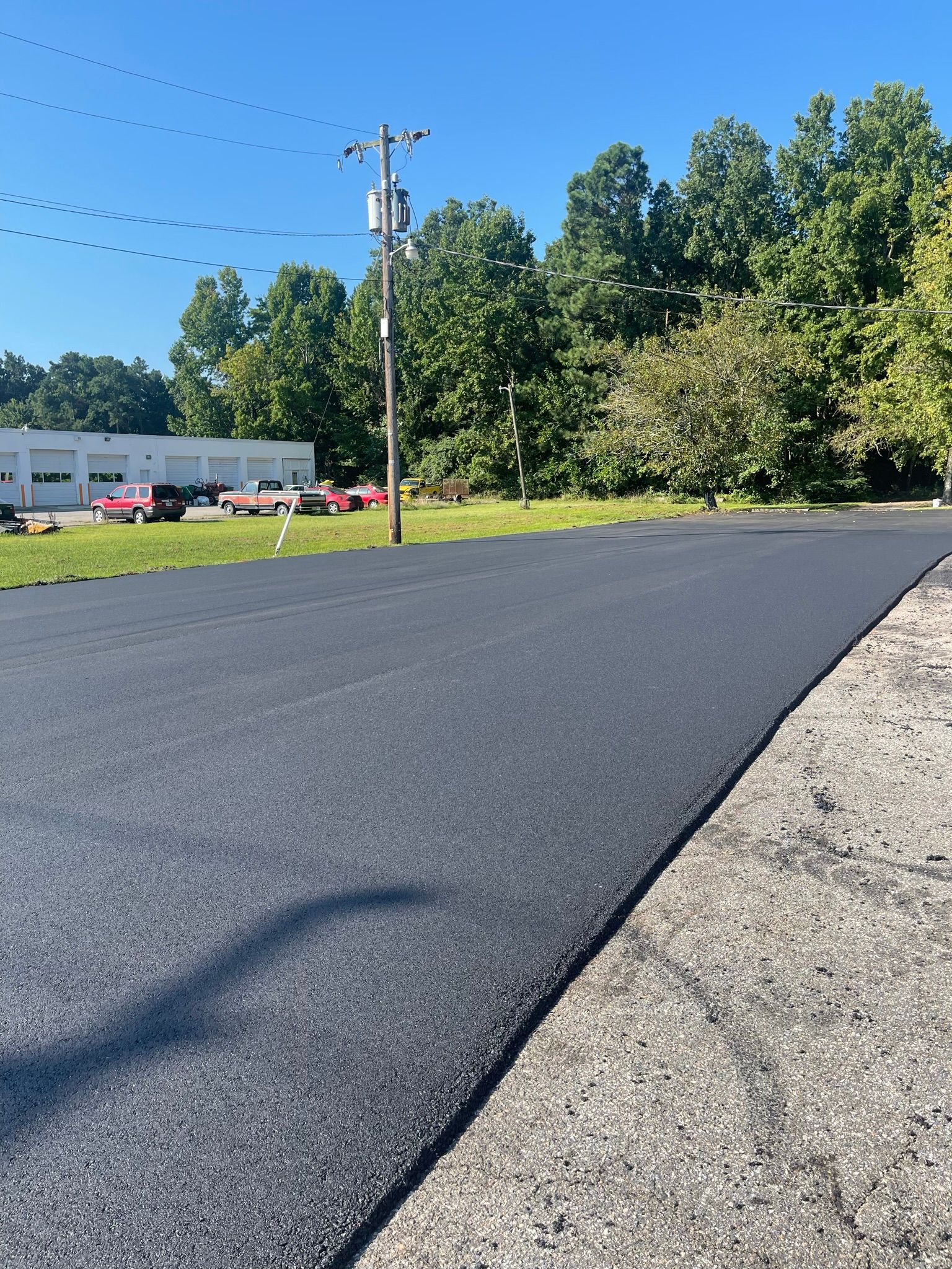 Freshly paved black road beside gravel shoulder, utility pole, and trees on a sunny day