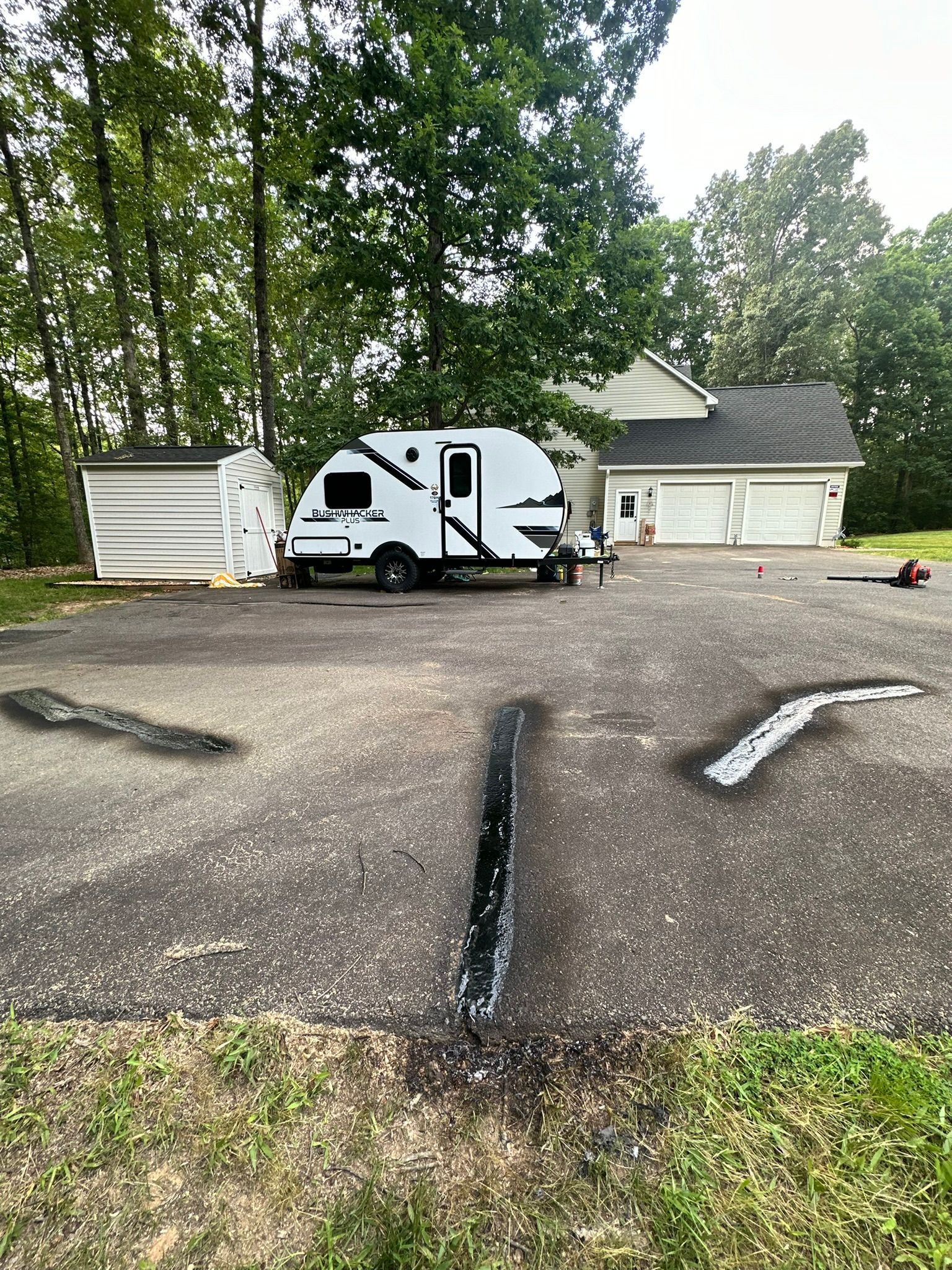 White RV parked in a wooded driveway beside a garage and white shed, with a cracked asphalt foreground