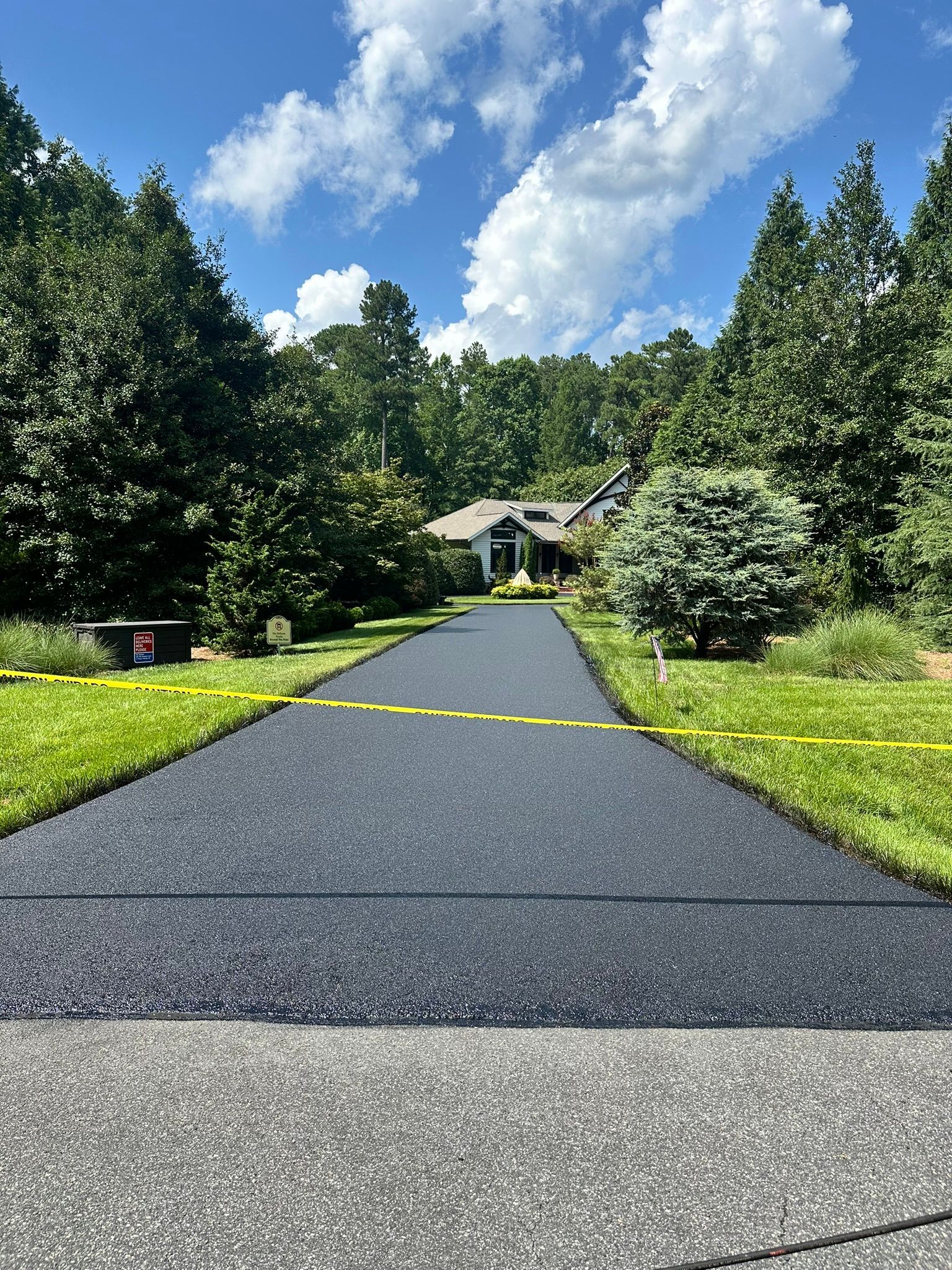 Tree-lined driveway leading to a house under a bright blue sky with white clouds.