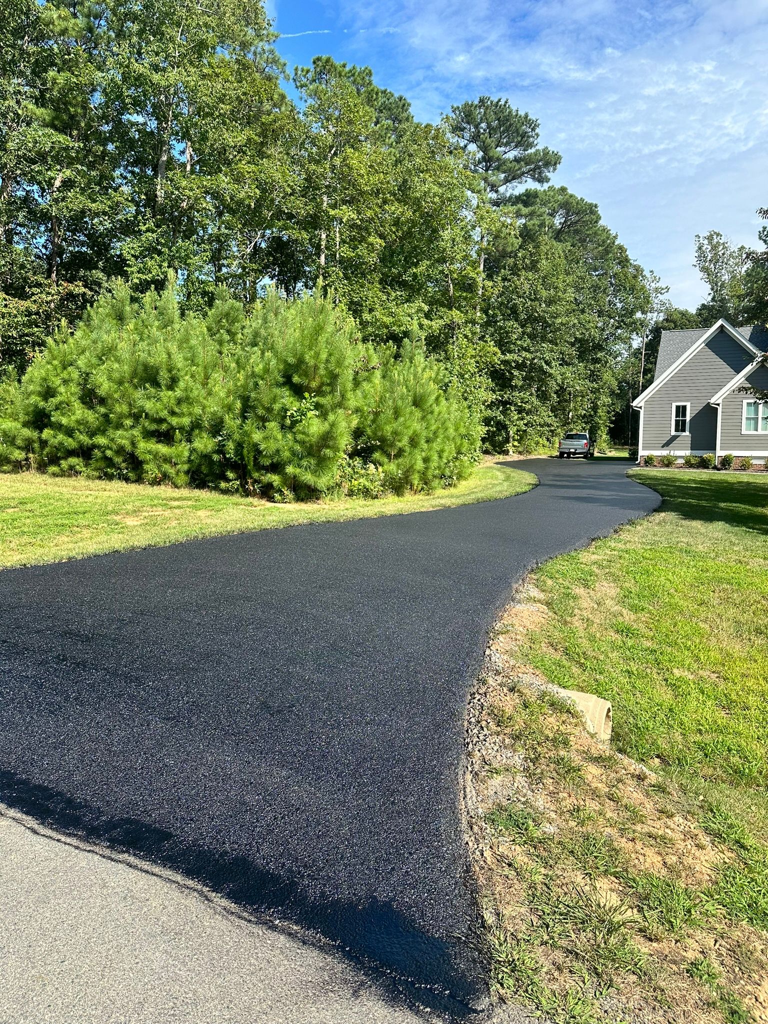 Freshly paved black driveway curving through trees beside a gray house on a sunny day