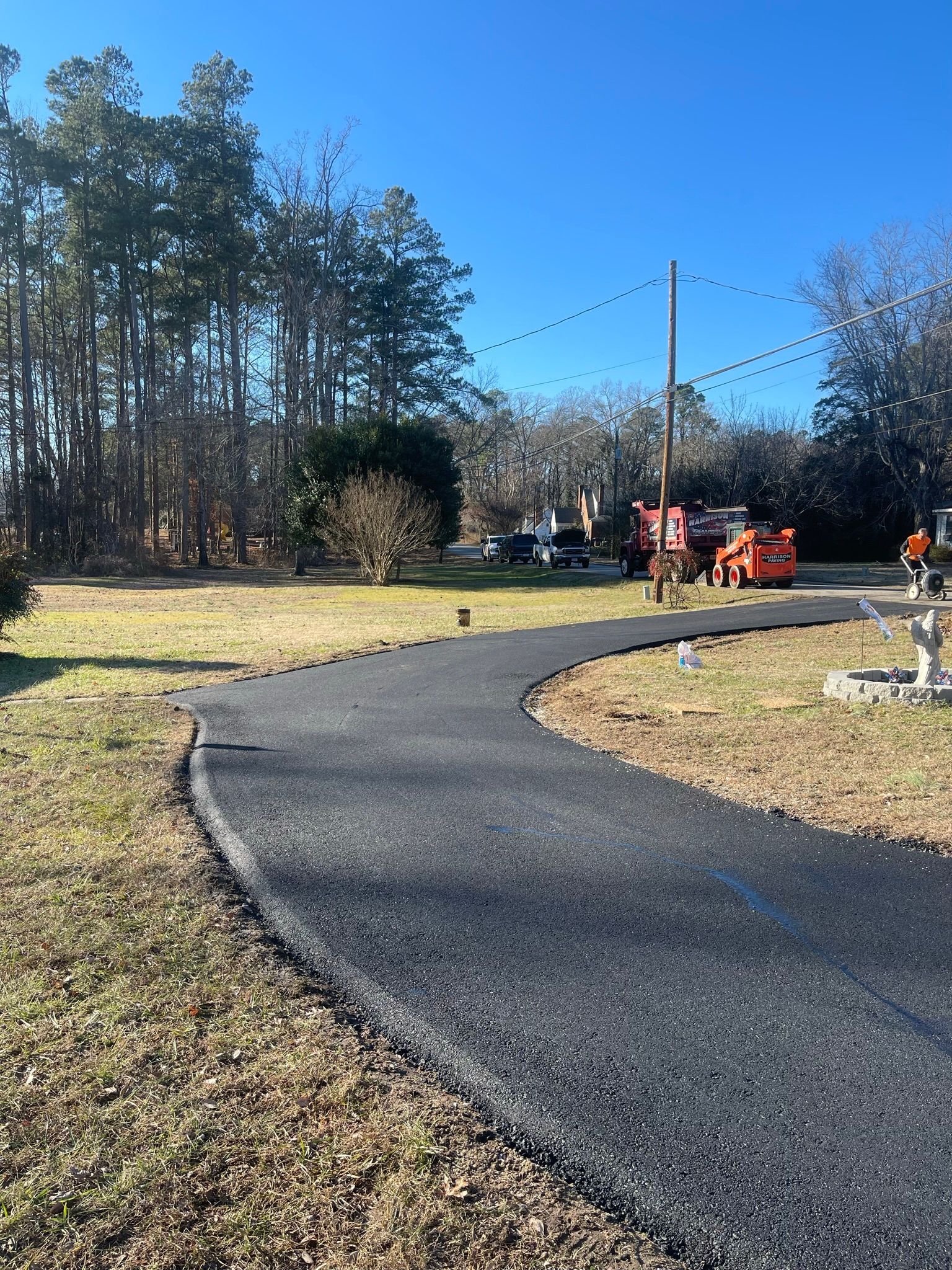 Curved black asphalt path beside a grassy yard and trees, with people and equipment in the distance under a clear sky
