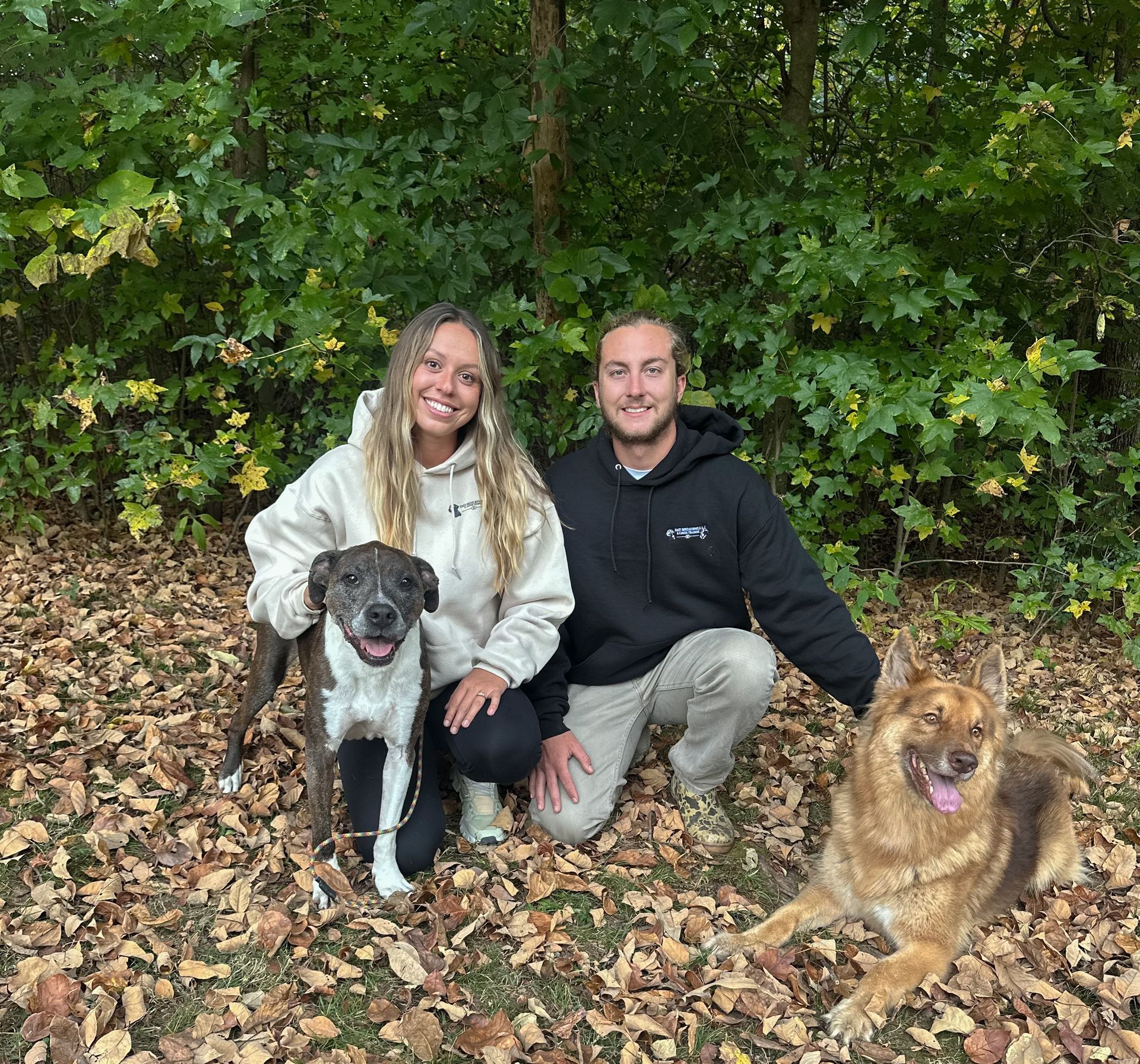 Couple kneels with two dogs in front of fall foliage. One dog is spotted, the other is brown.