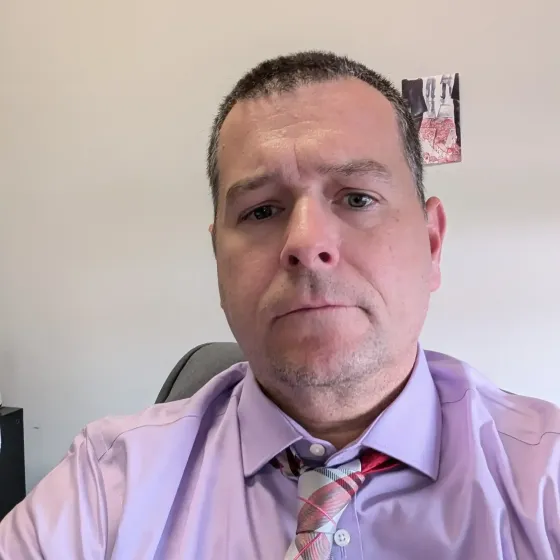 Person in a lavender shirt and patterned tie looking at the camera in an office.
